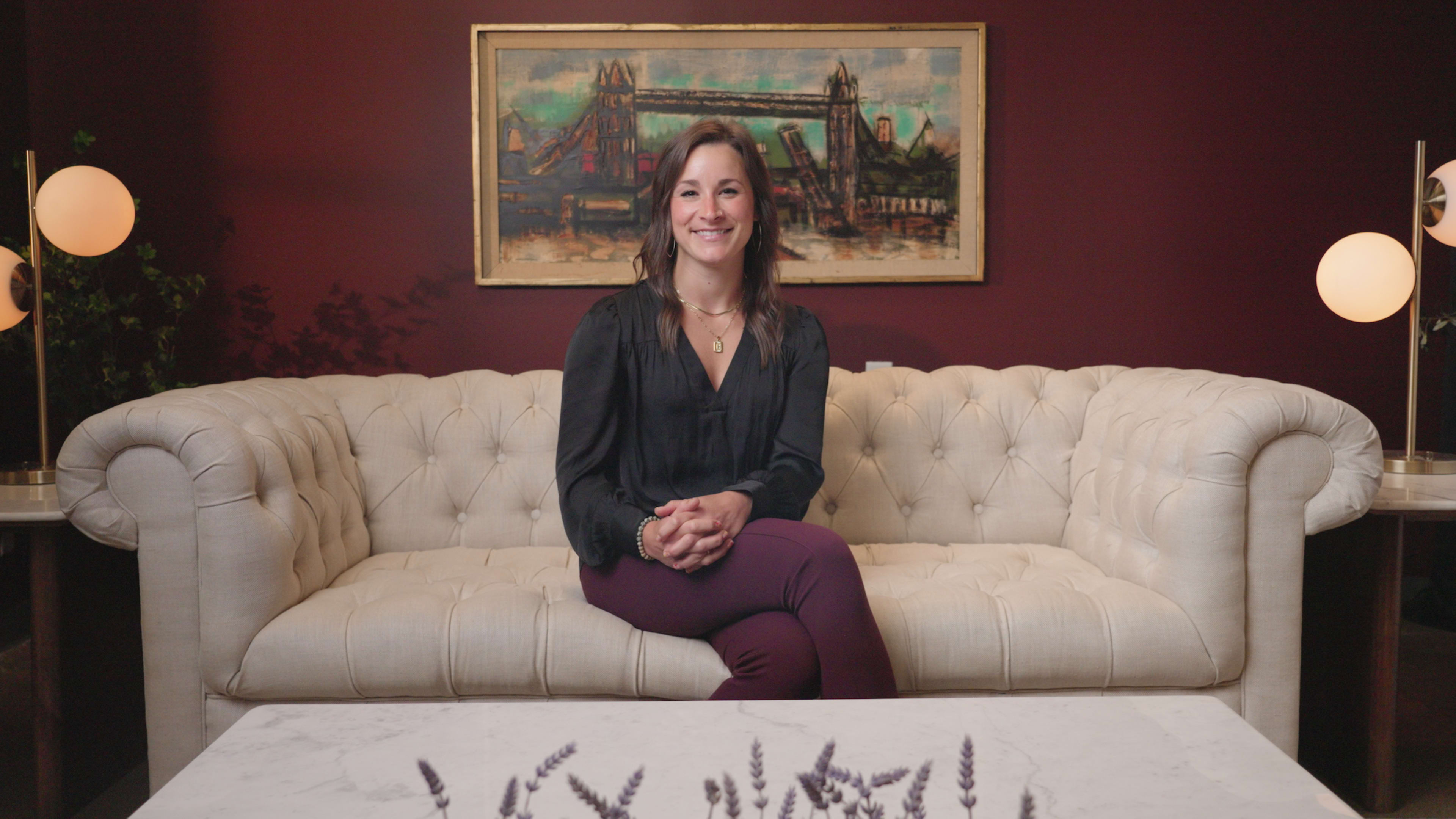 A woman sits on a tufted white sofa in a room with a dark red wall and a framed painting in the background.