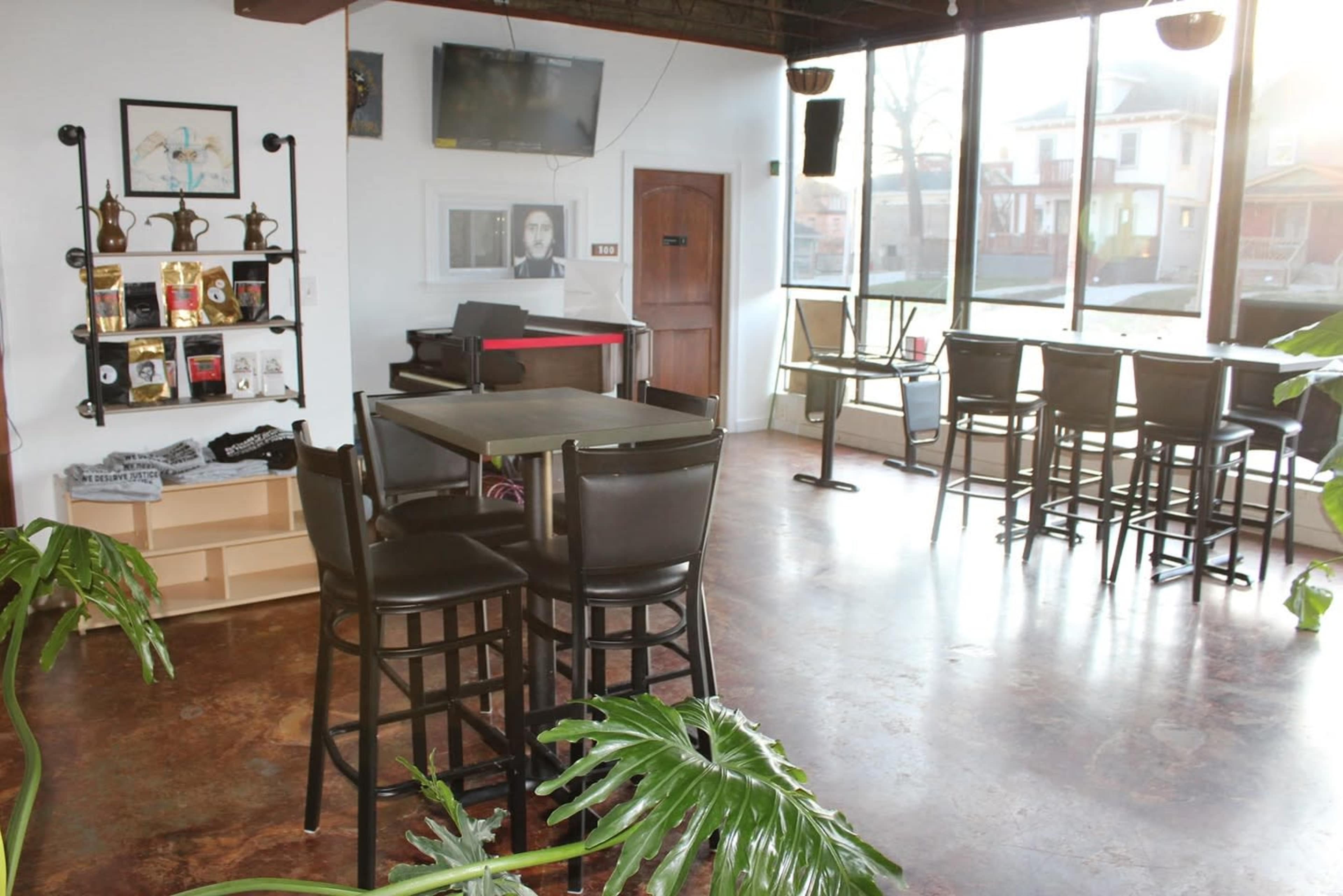 The image shows a modern cafe interior featuring tables and chairs, a bar top with high stools, and large windows allowing natural light to enter.