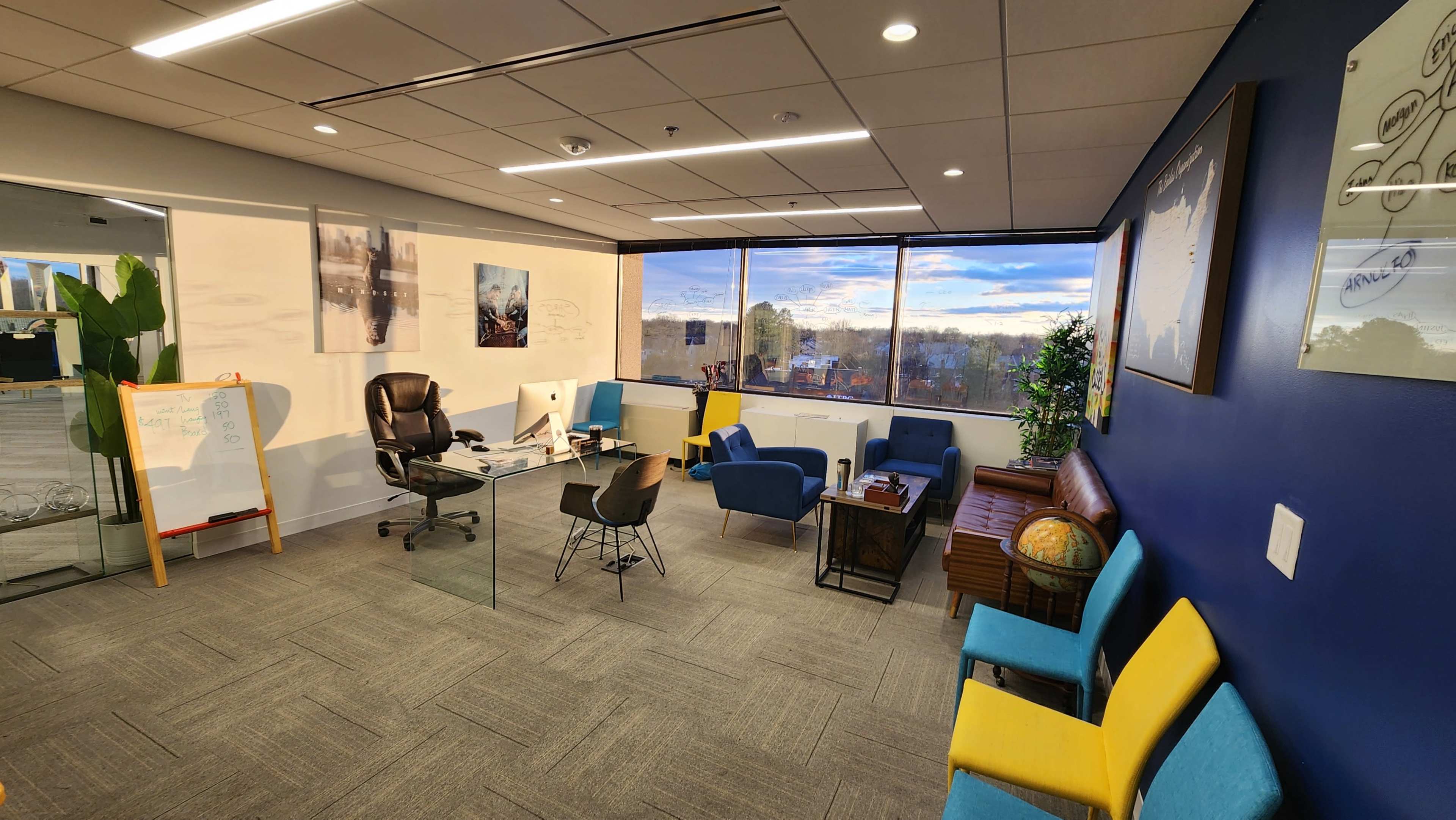 A modern office space featuring a glass desk with a black chair, various seating options in multiple colors, and large windows displaying a partially cloudy sky.