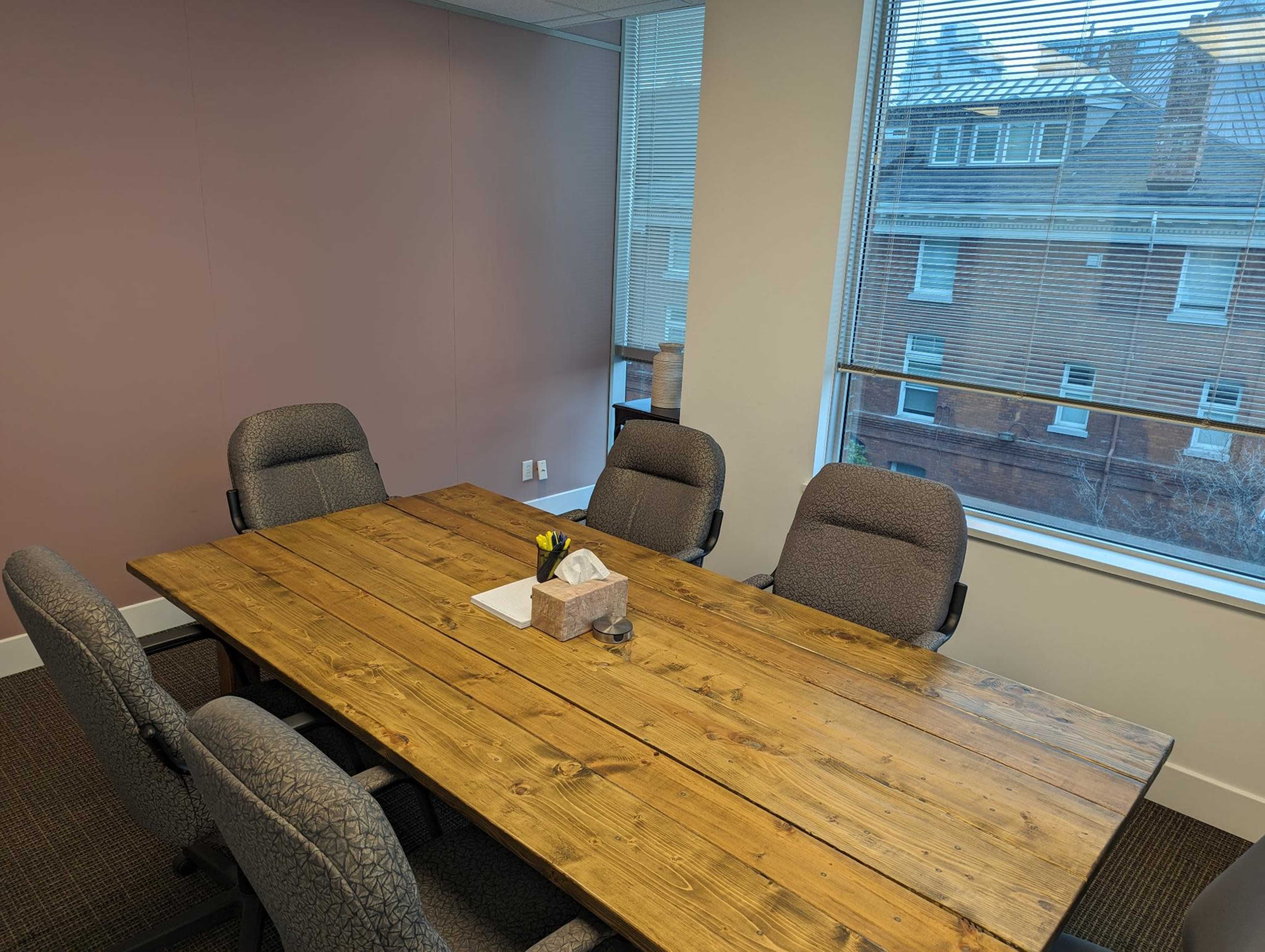 A rectangular wooden table is set with a notepad and a small decorative item in a conference room with gray chairs and large windows.