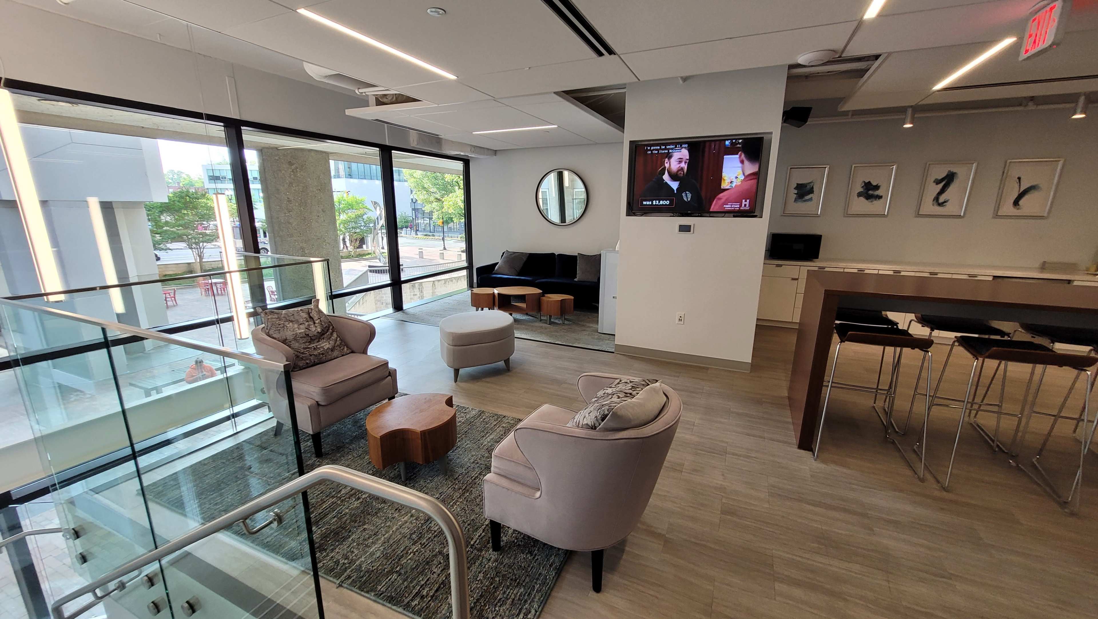 The image shows a modern lounge area with comfortable seating, a television, and large windows letting in natural light.