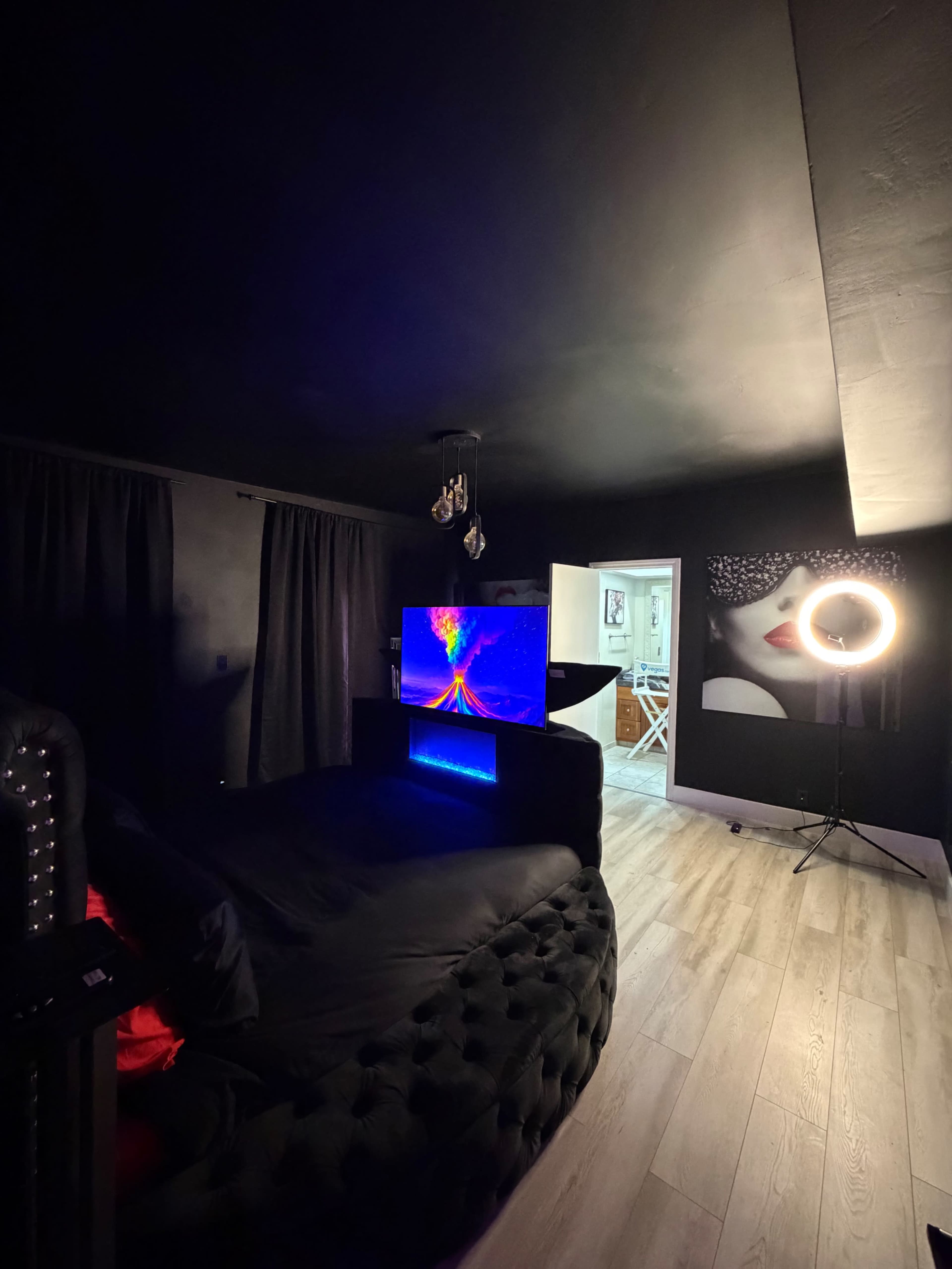 The image depicts a dark bedroom featuring a large black tufted bed, a colorful television display, a ring light, and an adjoining room visible in the background.