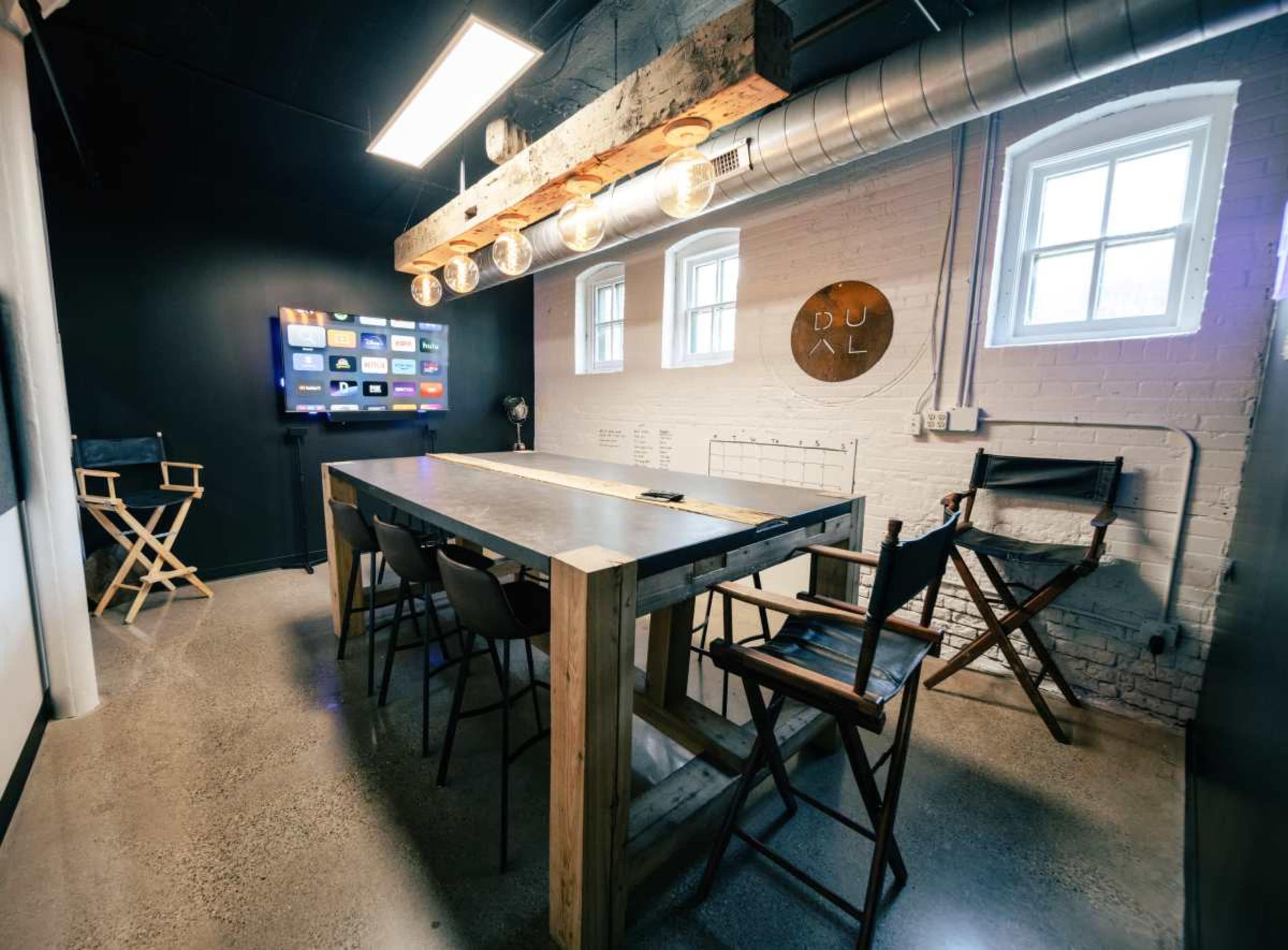 A modern meeting room featuring a long wooden table with high chairs, a wall-mounted screen, and exposed lighting.