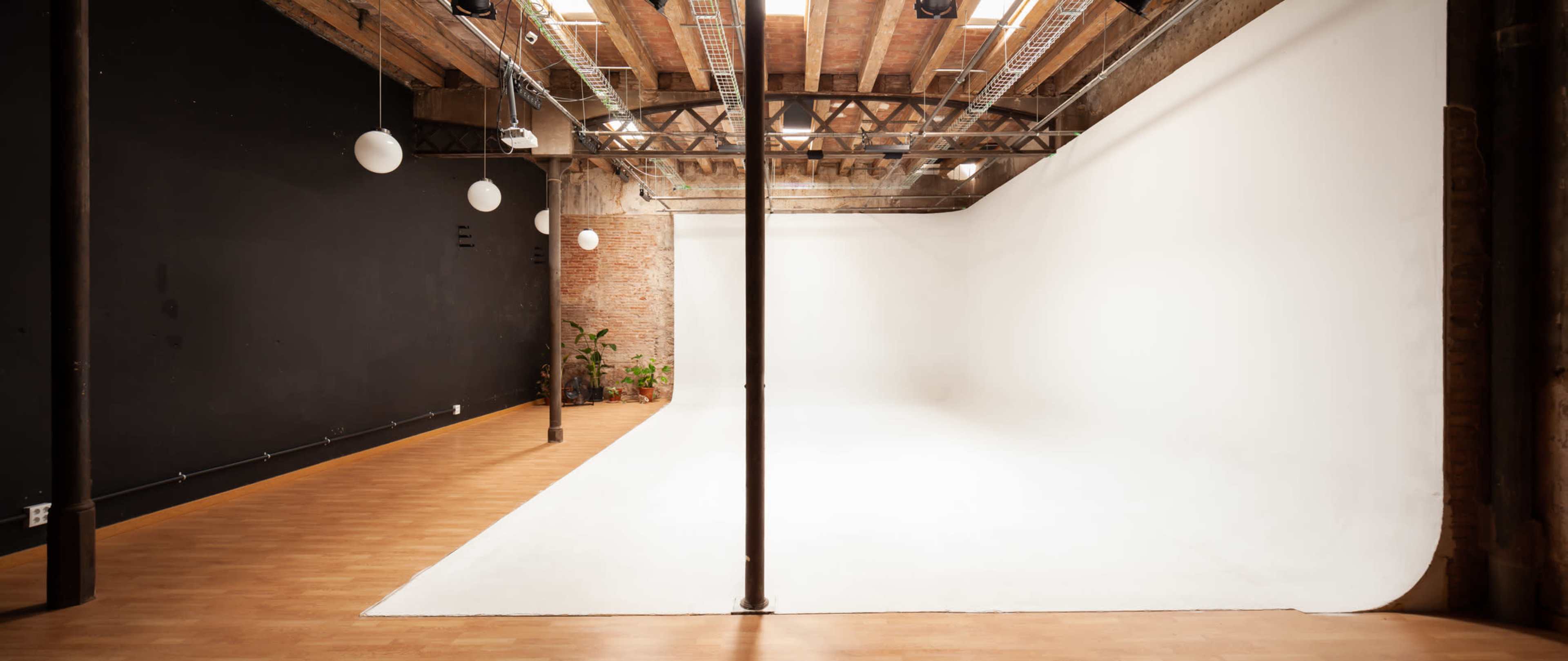 A spacious studio with a black-painted wall on one side and a white backdrop on the other, complemented by wooden floors and hanging light fixtures.