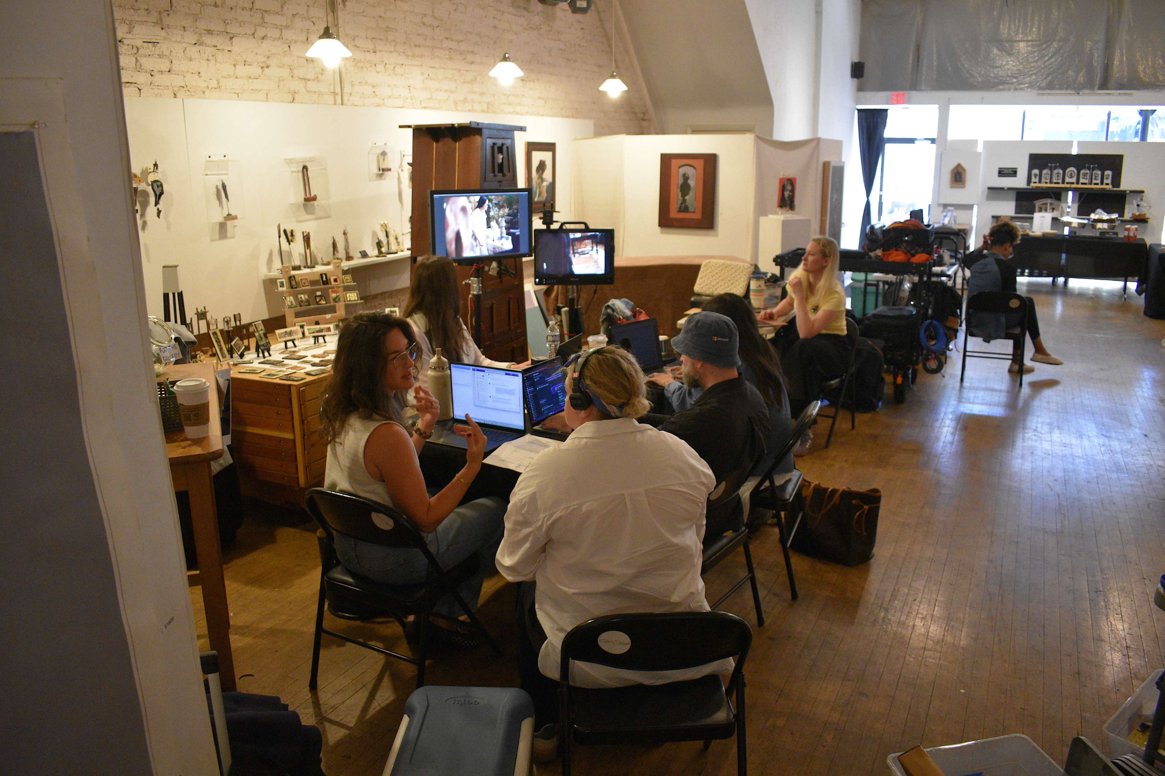 A group of people is working in a studio, seated around tables with computers and equipment, while others are positioned in the background among various artworks and supplies.