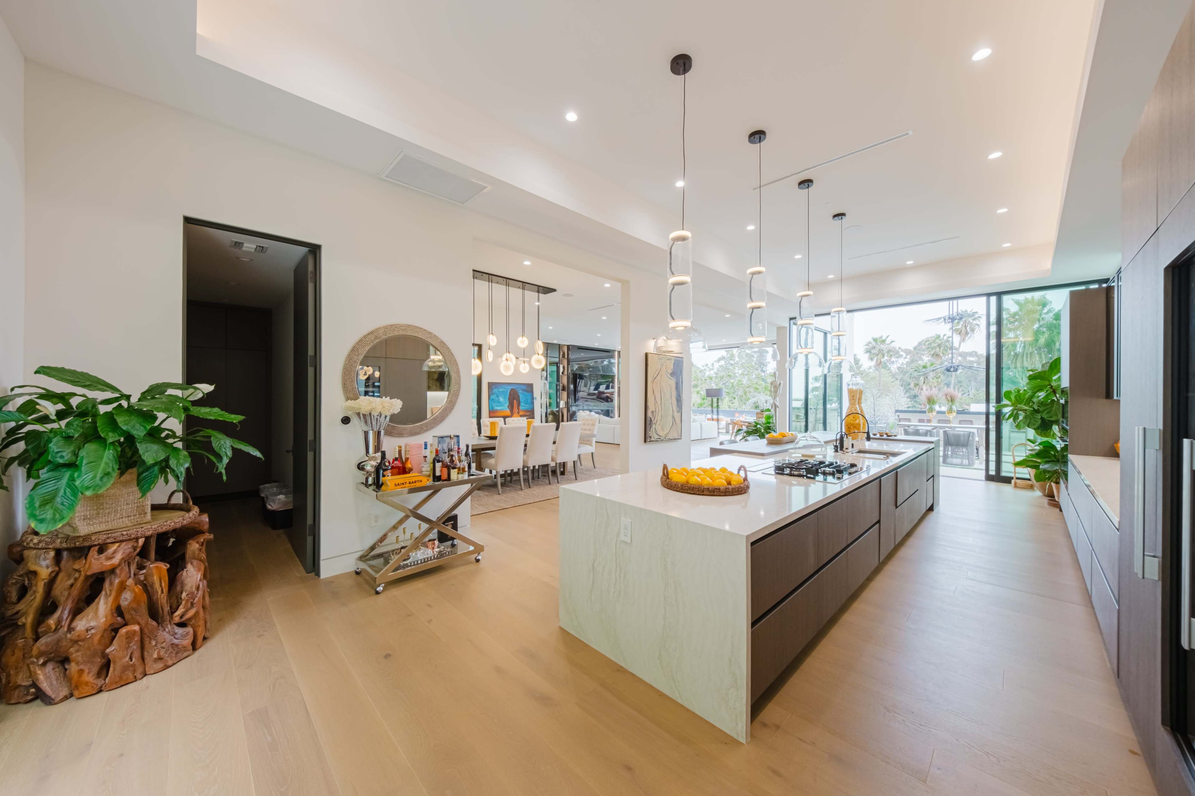 A modern kitchen features a large island with a countertop, pendant lights, and an open layout that connects to a dining area and outdoor space.