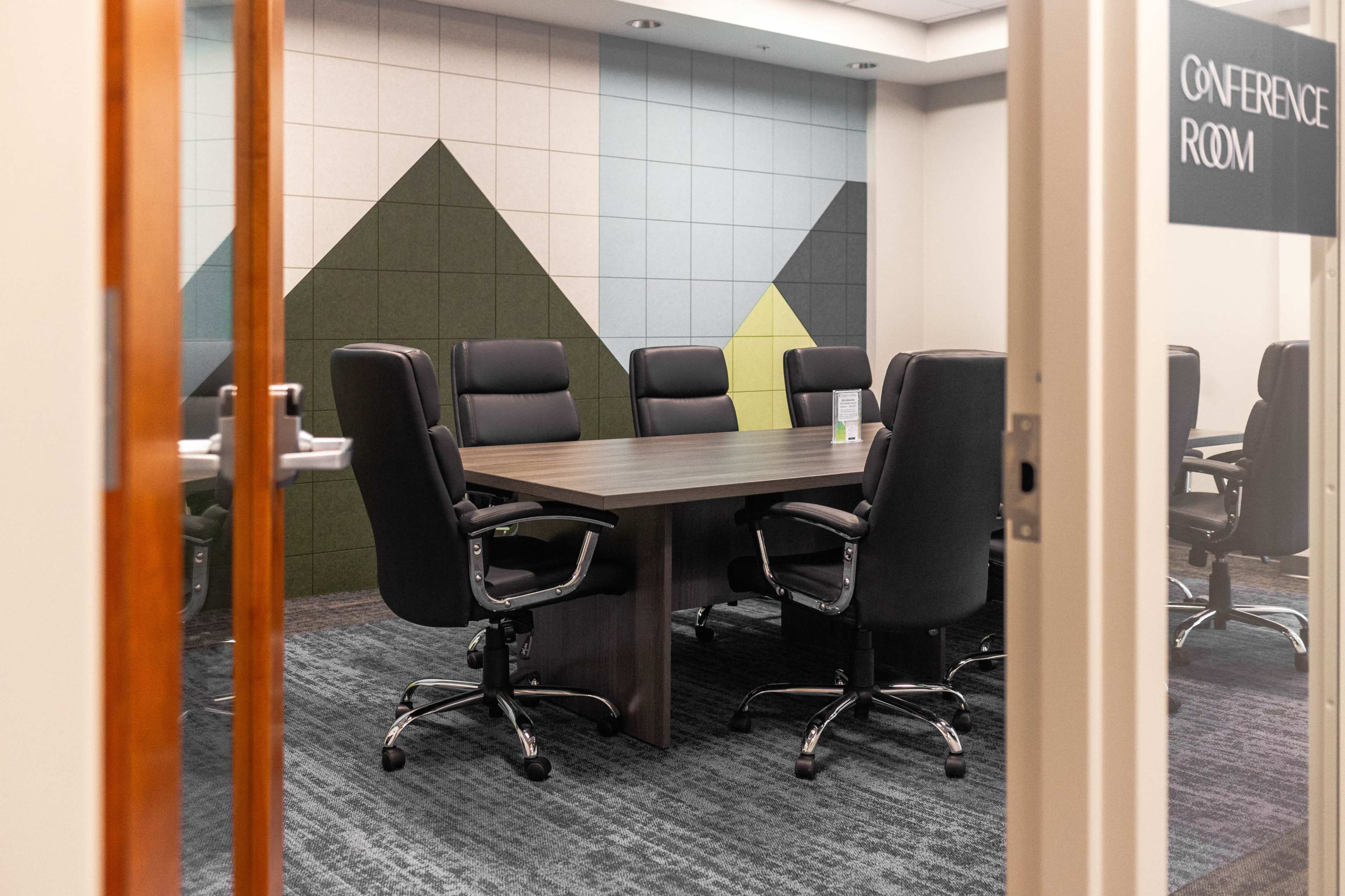A conference room features a large table surrounded by black rolling chairs, set against a colorful geometric wall.