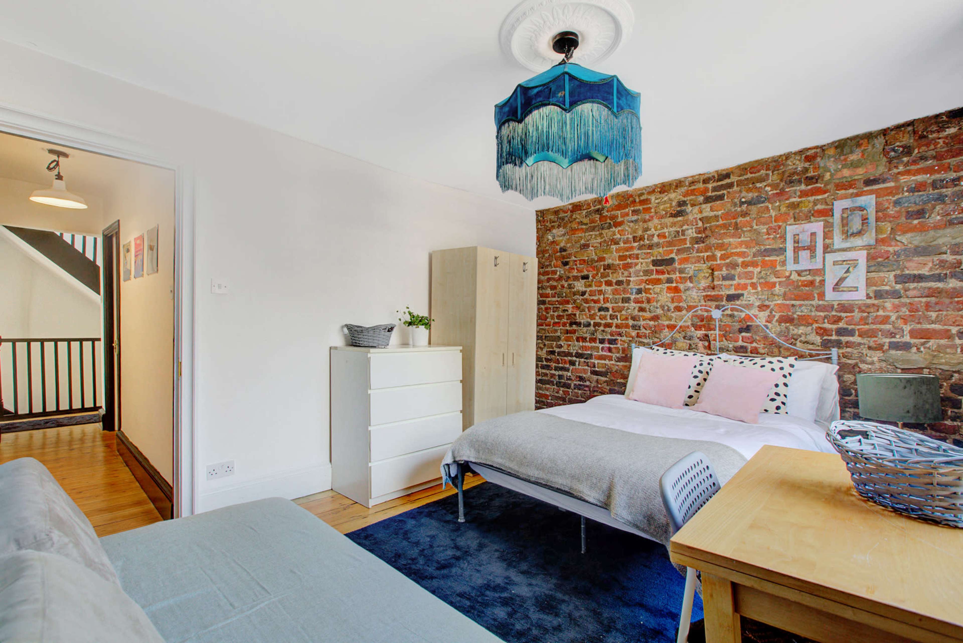 The image shows a bedroom featuring a brick accent wall, a bed with light bedding, a wooden table, and a wardrobe, illuminated by a colorful ceiling light.
