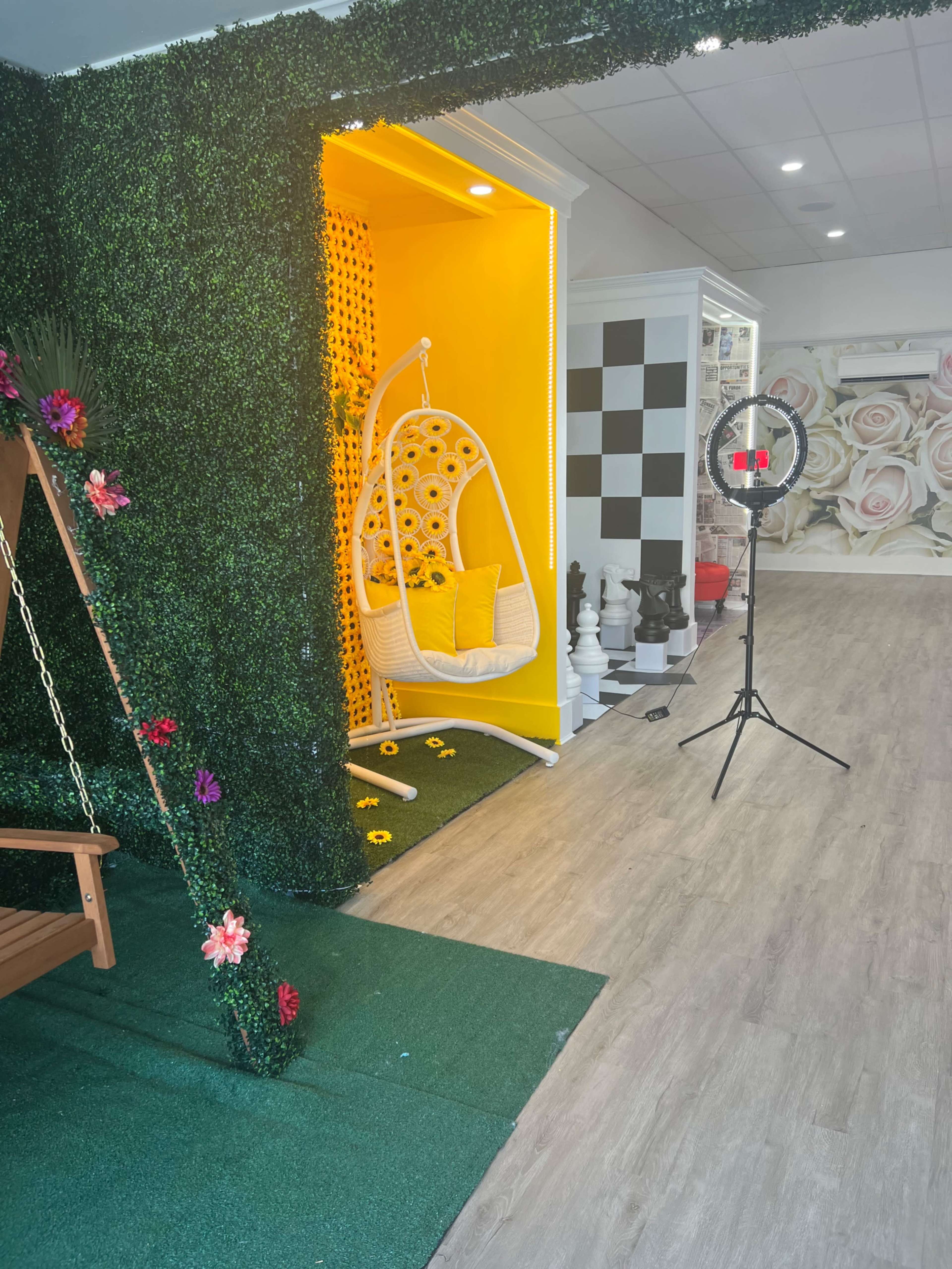 A brightly colored interior space featuring a yellow hanging chair surrounded by artificial greenery and a backdrop of pastel floral wallpaper.