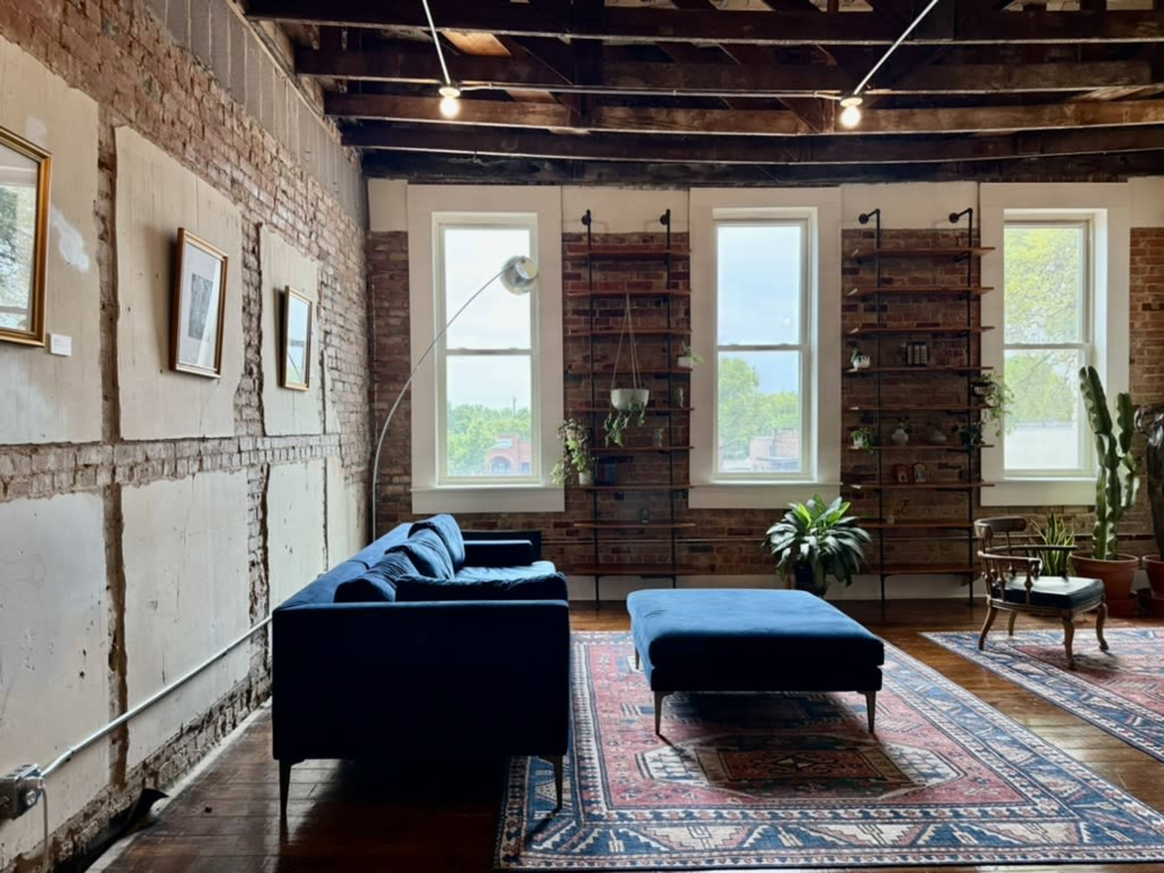 A stylish living room features a blue sofa and ottoman on a patterned rug, accented by large windows and exposed brick walls.