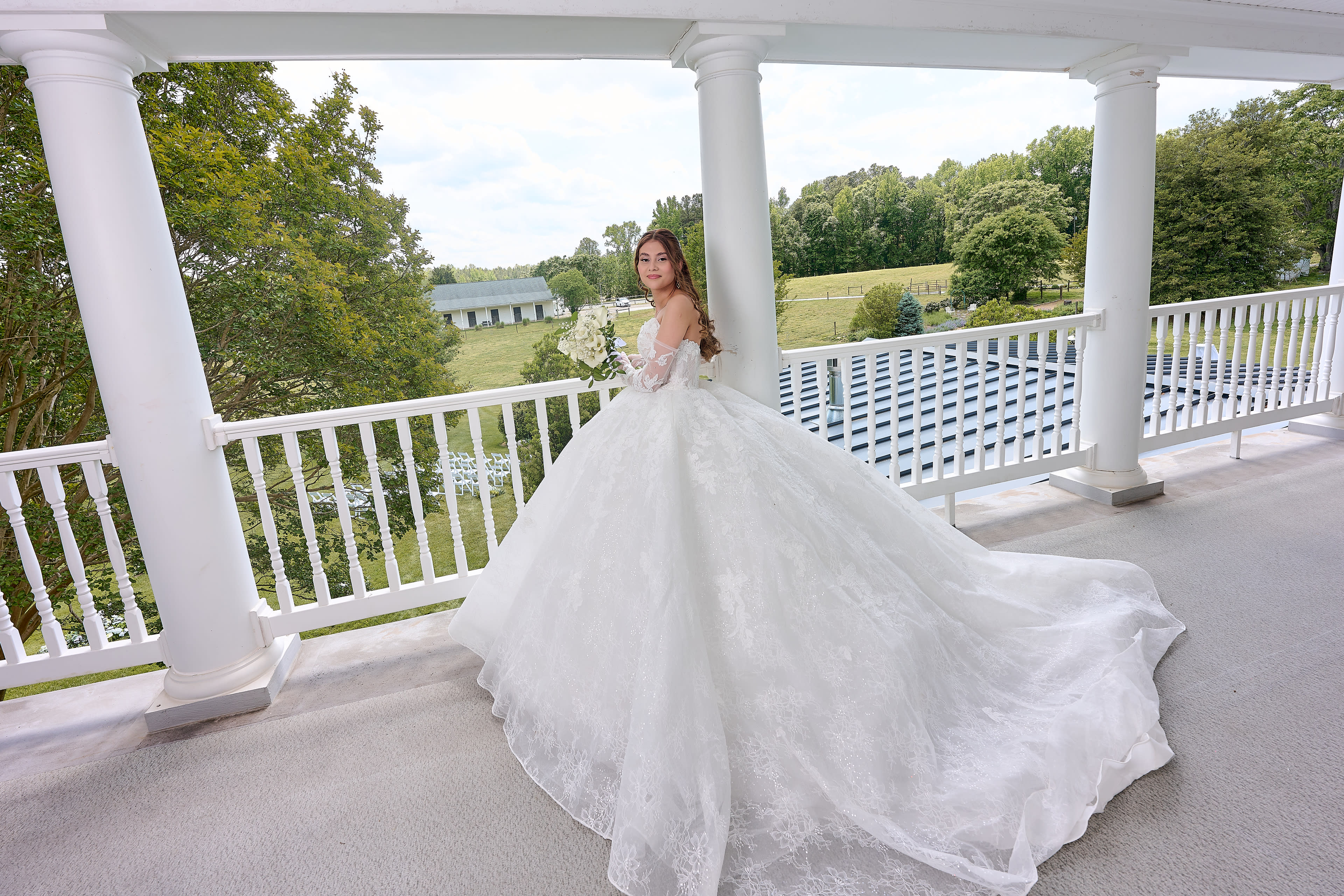 A woman in a white lace wedding dress stands on a balcony overlooking a landscaped area with seating arranged for an event.