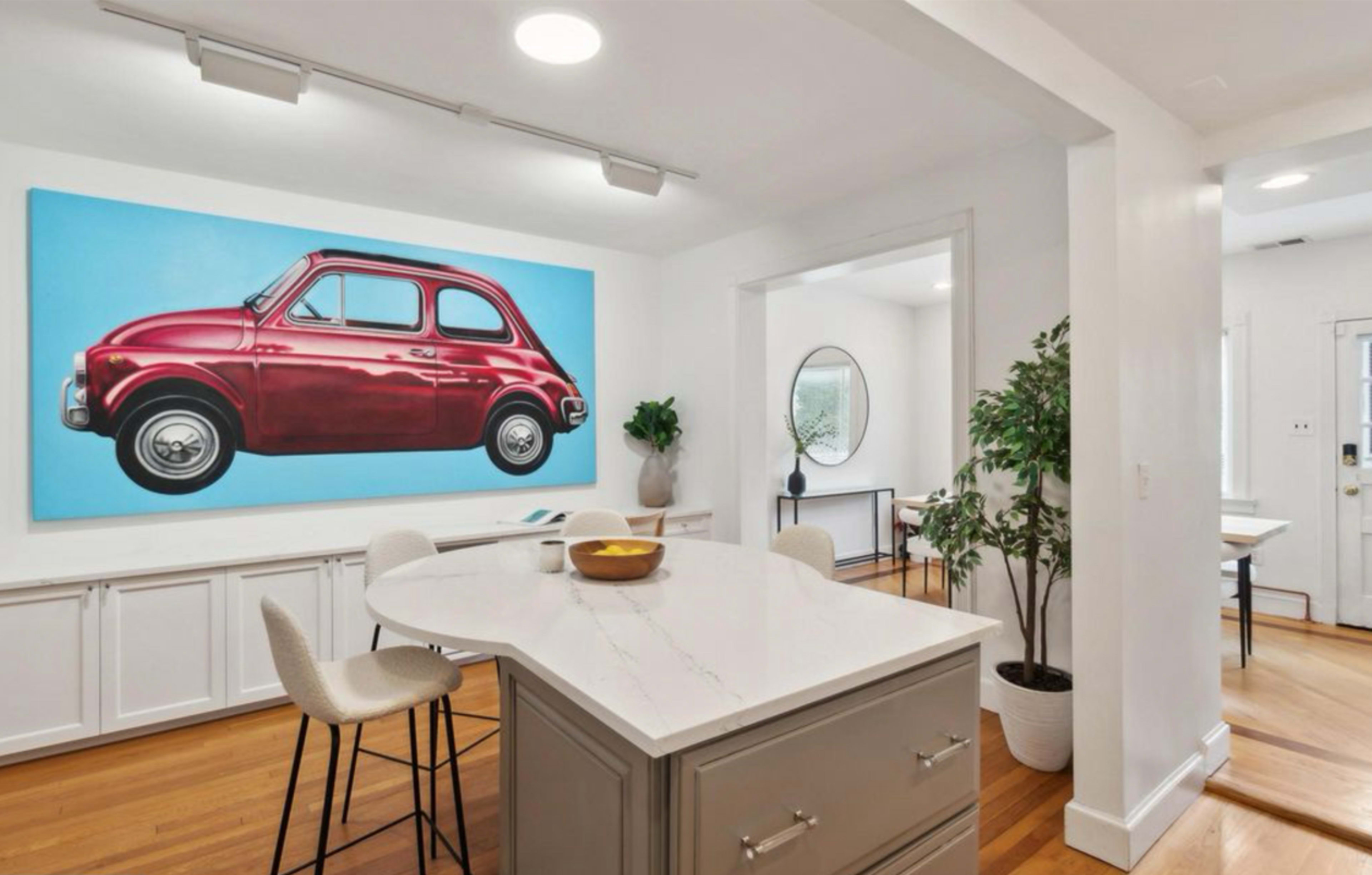 A red vintage car is depicted on a large canvas hanging on the white wall of a modern kitchen with a circular dining table and wooden floors.
