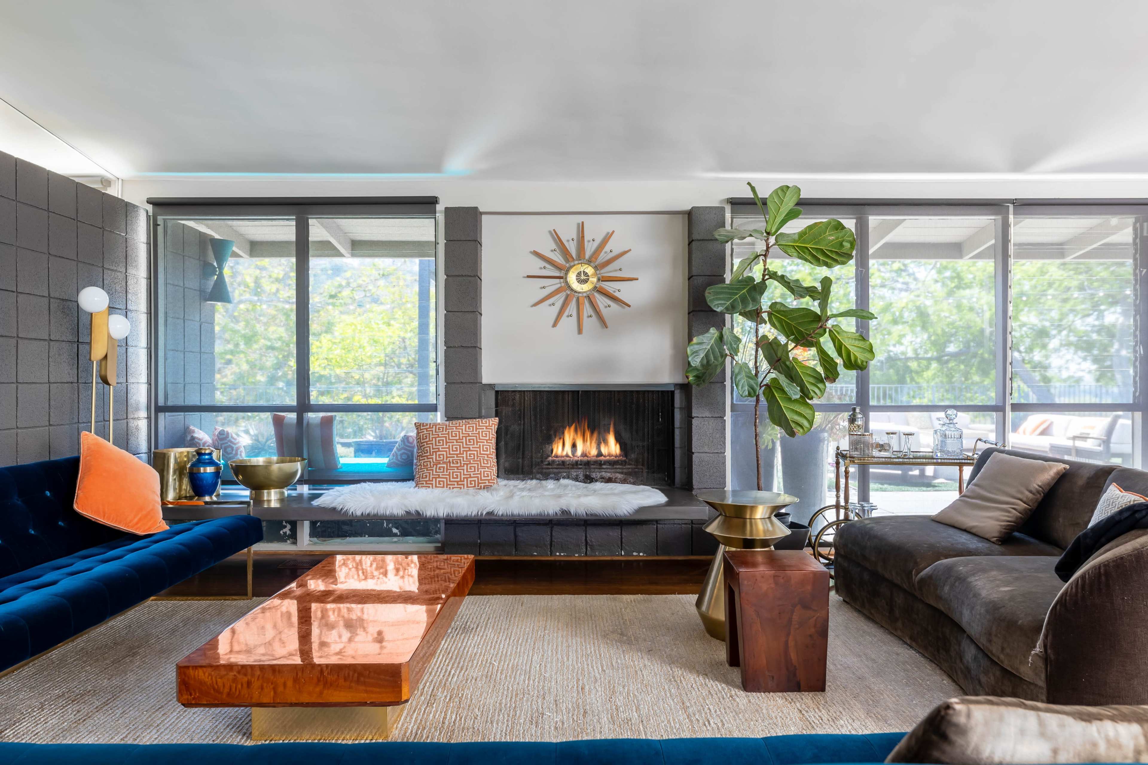 A modern living room features a fireplace, large windows, a blue velvet sofa, and a wooden coffee table.