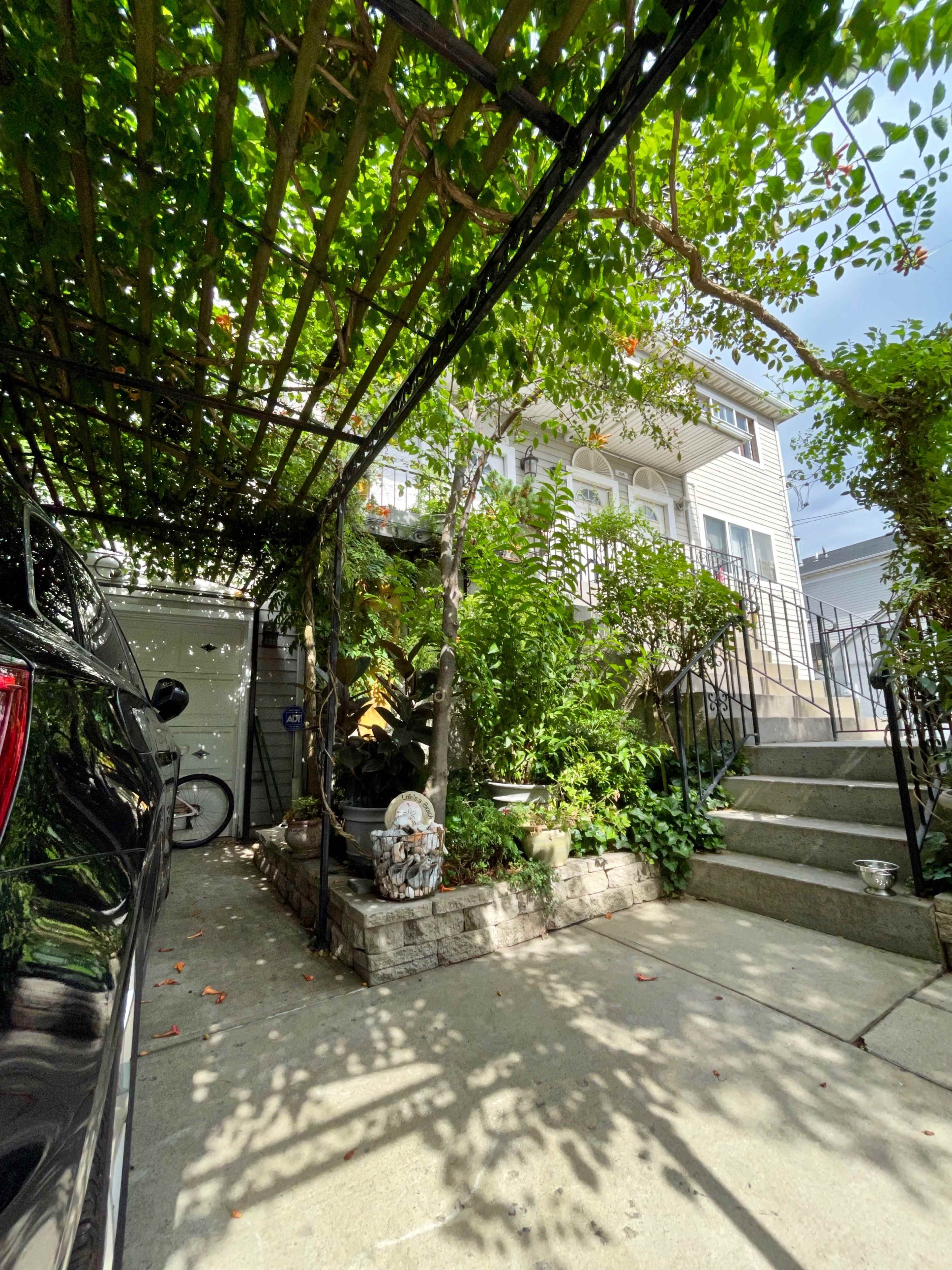 A car is parked under a trellis covered with vines, leading to a yard with steps and a well-maintained garden beside a house.