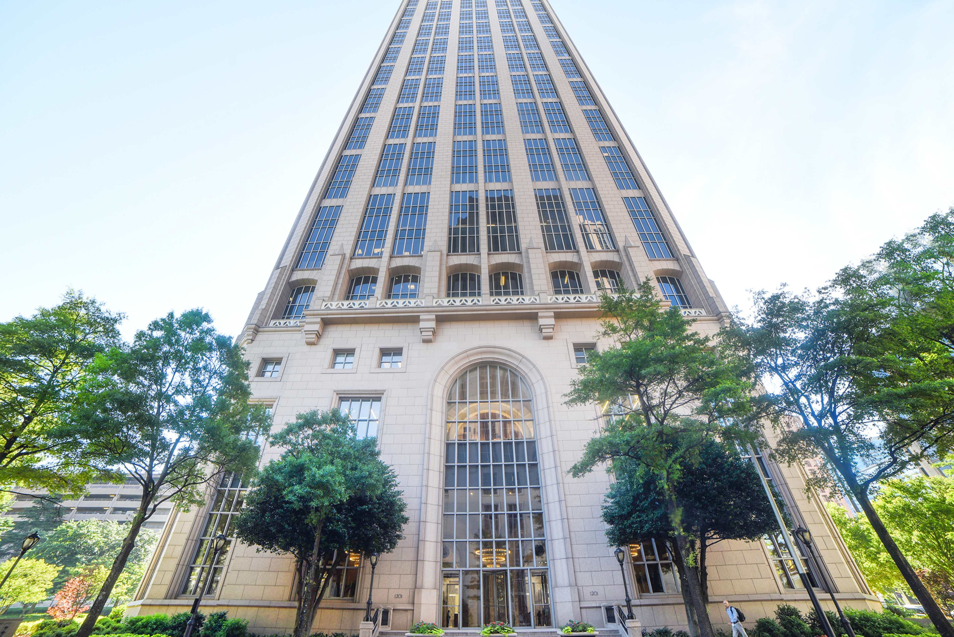 The image shows a tall, modern skyscraper with a grand entrance, framed by trees in the foreground.
