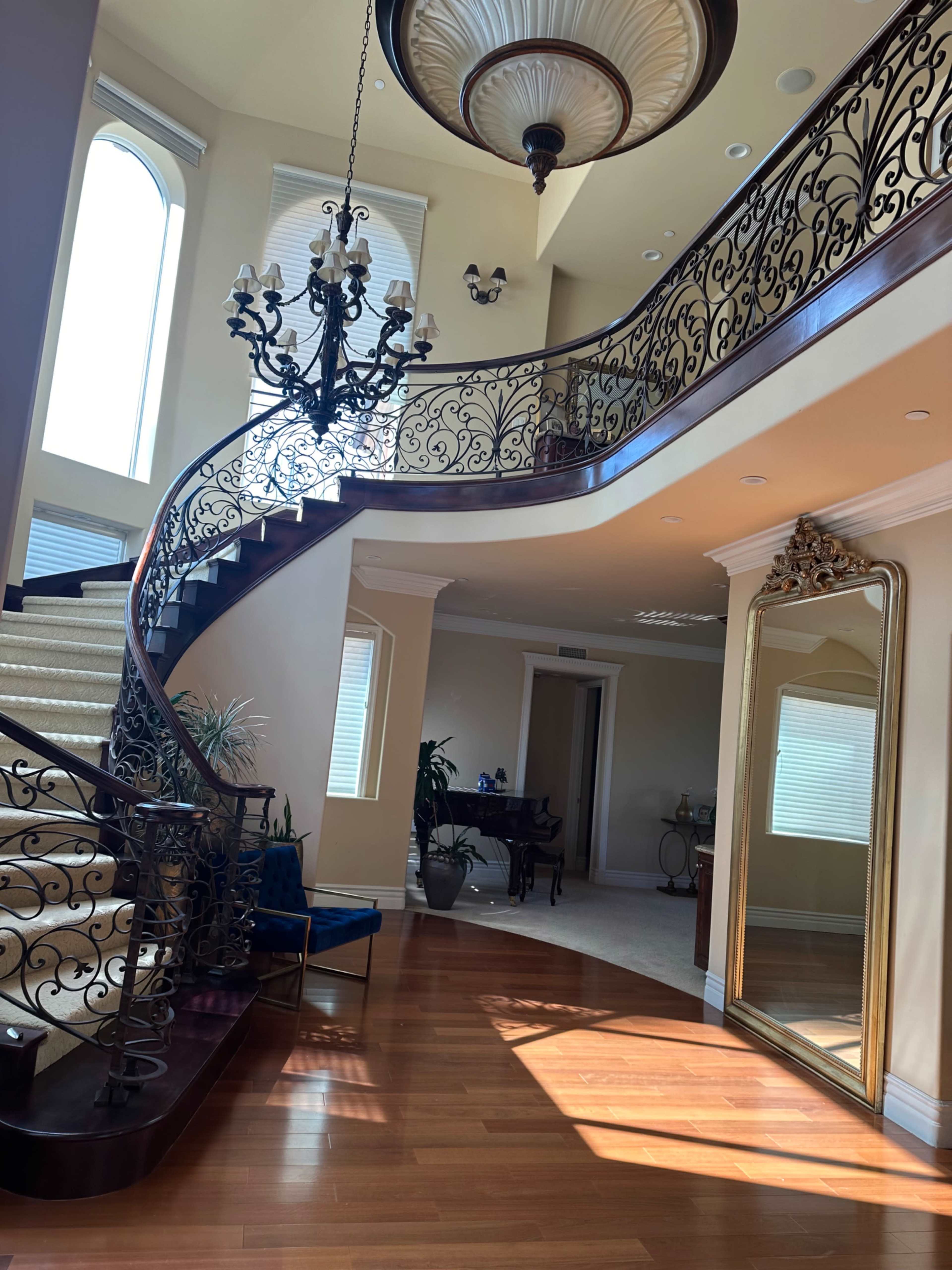 The image depicts a grand foyer with a curved staircase, ornate ironwork railing, and large windows, illuminated by sunlight.