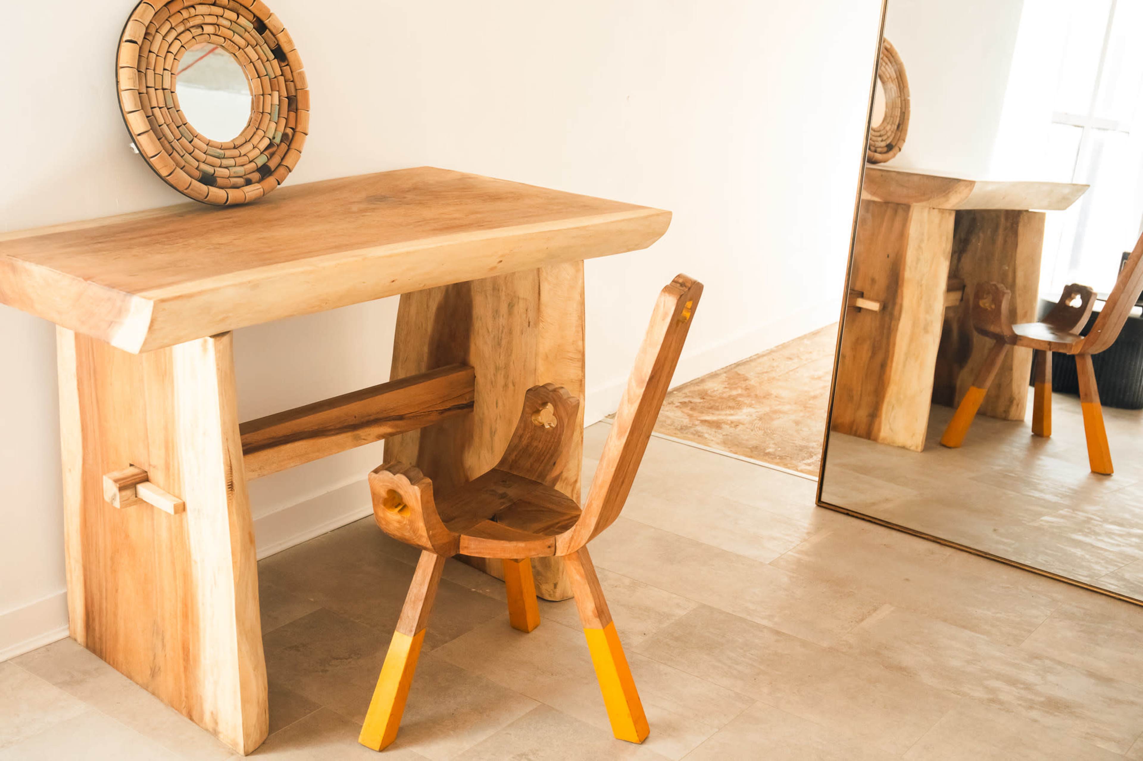 A wooden desk with a matching chair featuring yellow legs is positioned next to a large mirror and a round decorative wall piece.