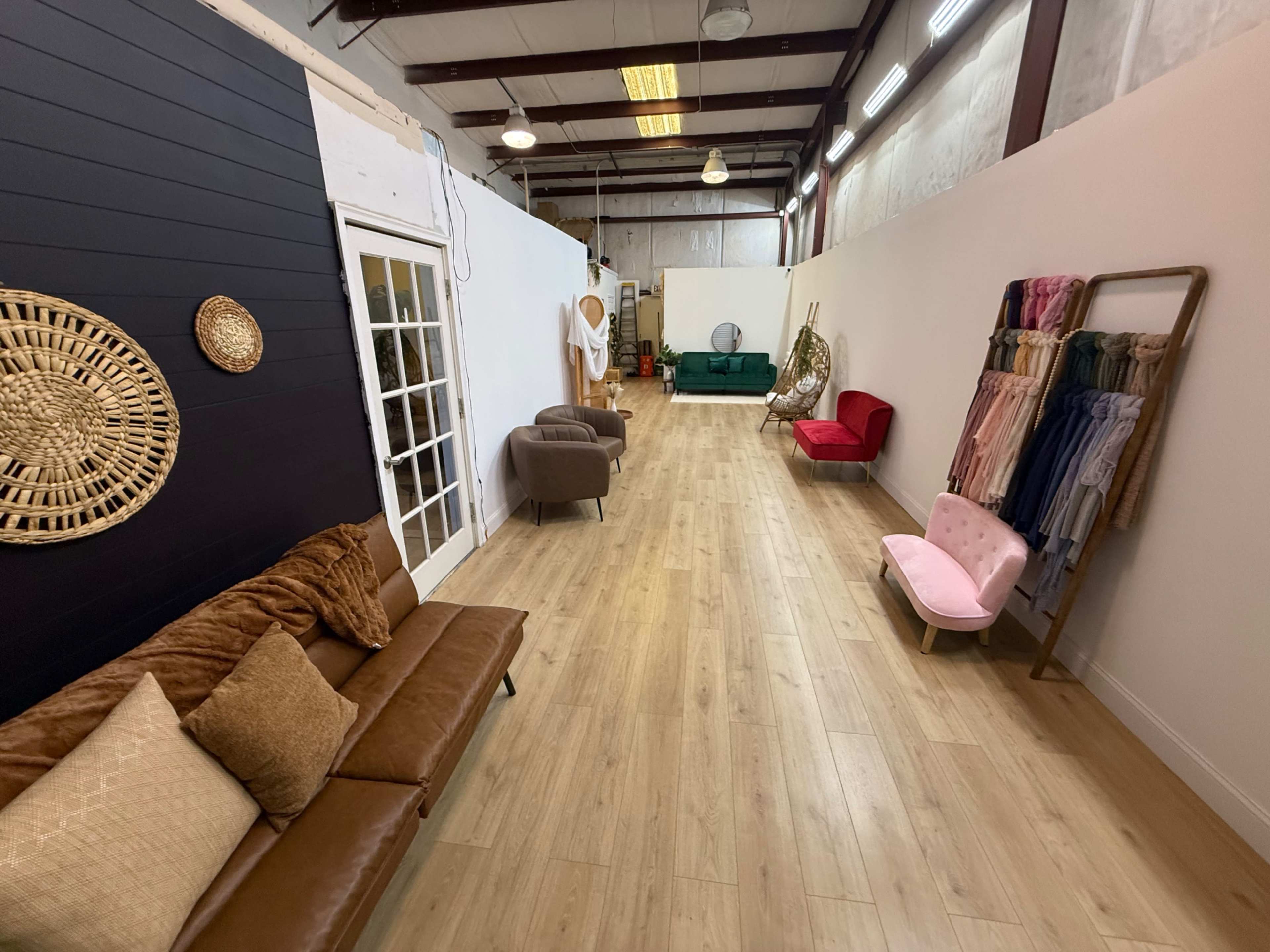 The image shows a spacious interior with a wooden floor, featuring a brown couch, chairs, and various decorative textiles displayed on a rack against a white wall.