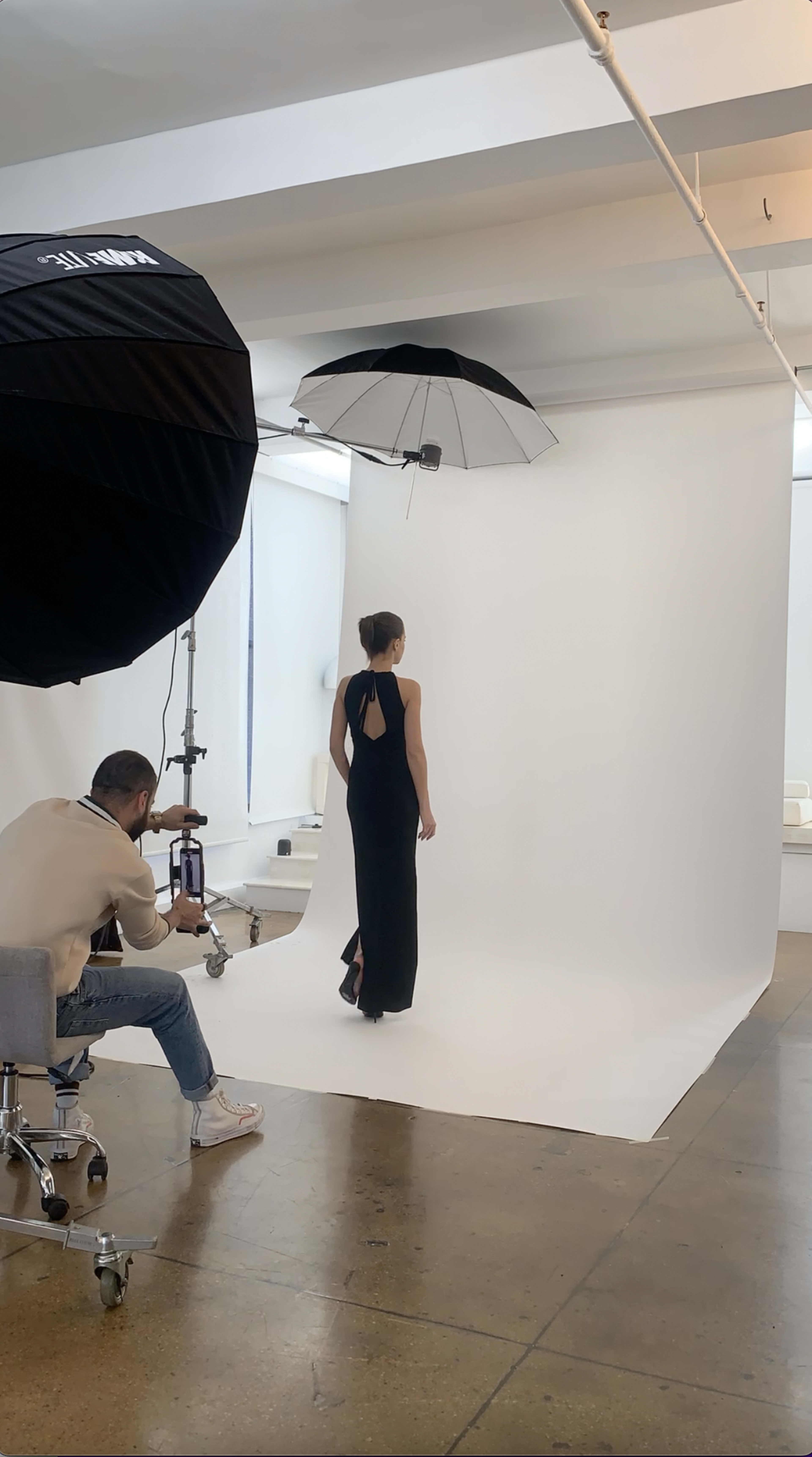 A model wearing a black dress poses under an umbrella in a photography studio, while the photographer adjusts his camera.