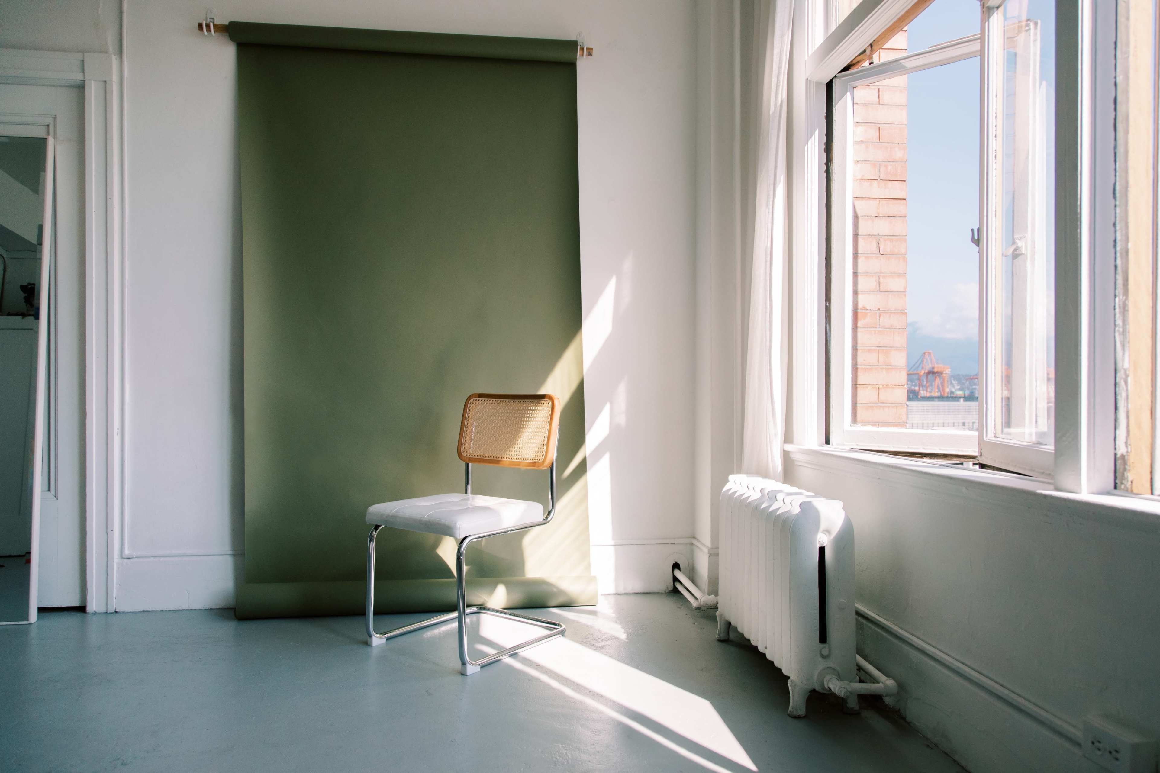 A minimalist room features a single chair in front of a green backdrop, with sunlight streaming in through a window.