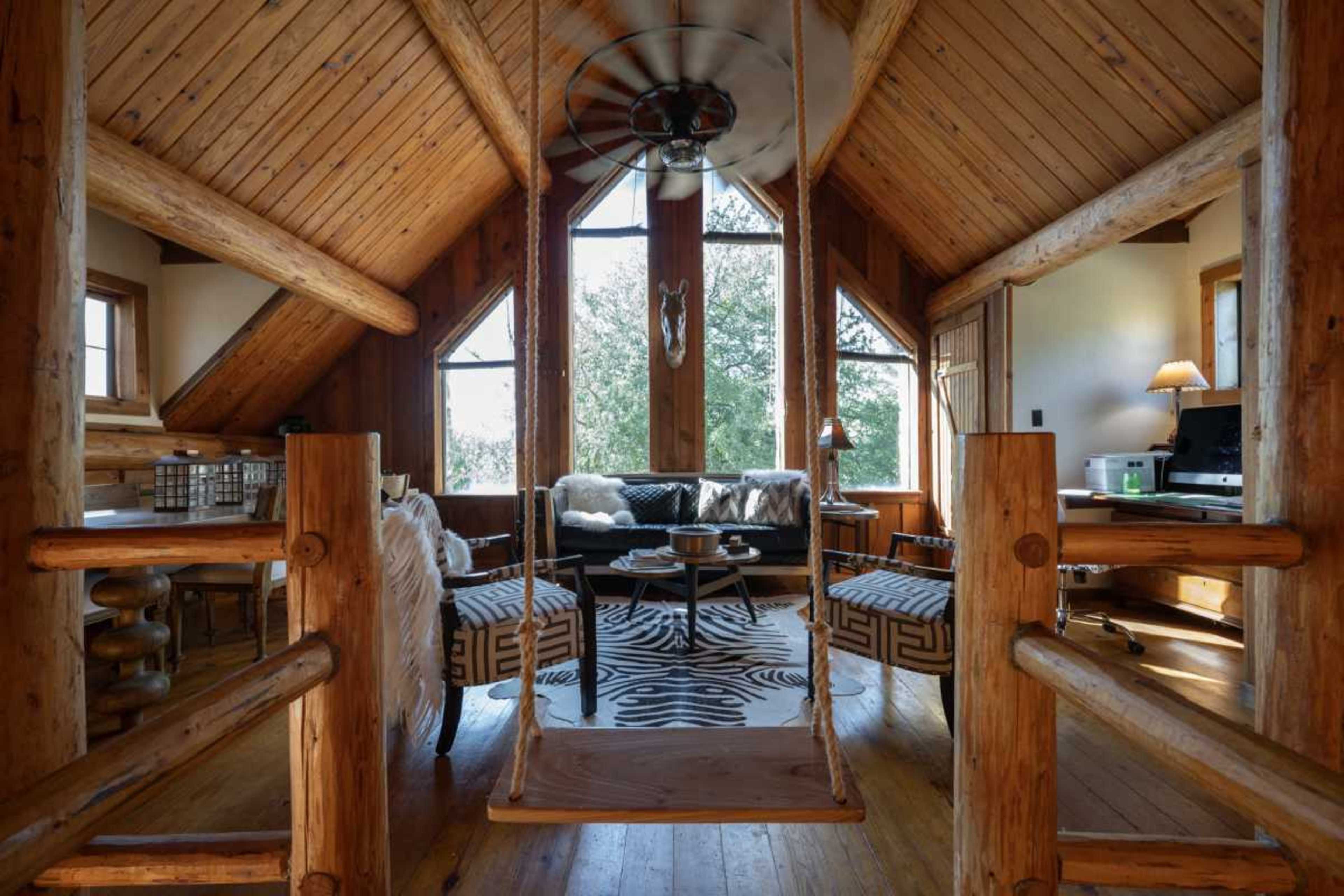 A wooden cabin interior features a swing and a cozy seating area with a view of the outdoors through large windows.