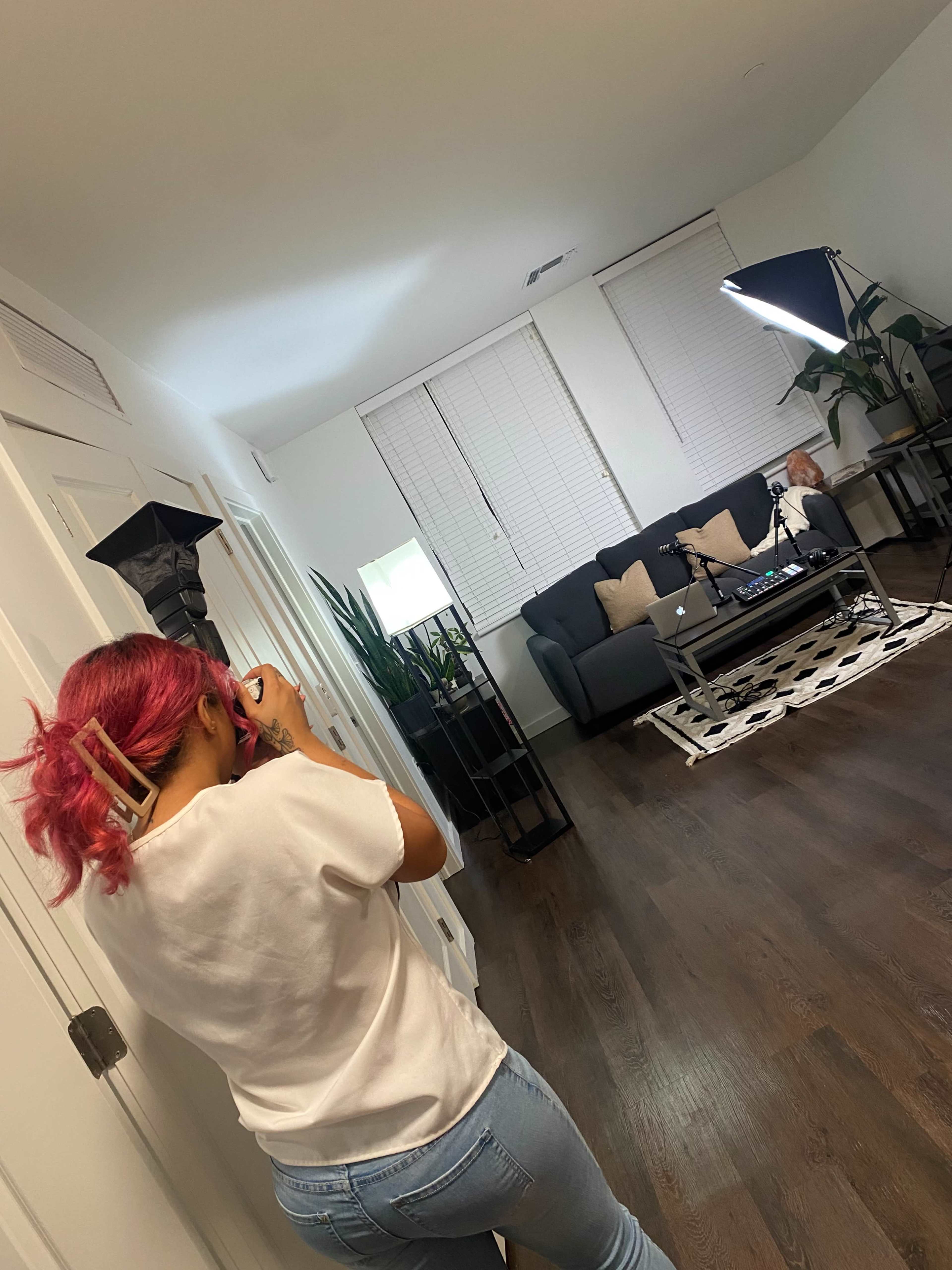A person with pink hair takes a photo in a well-lit living room featuring a sofa, coffee table, and plants.