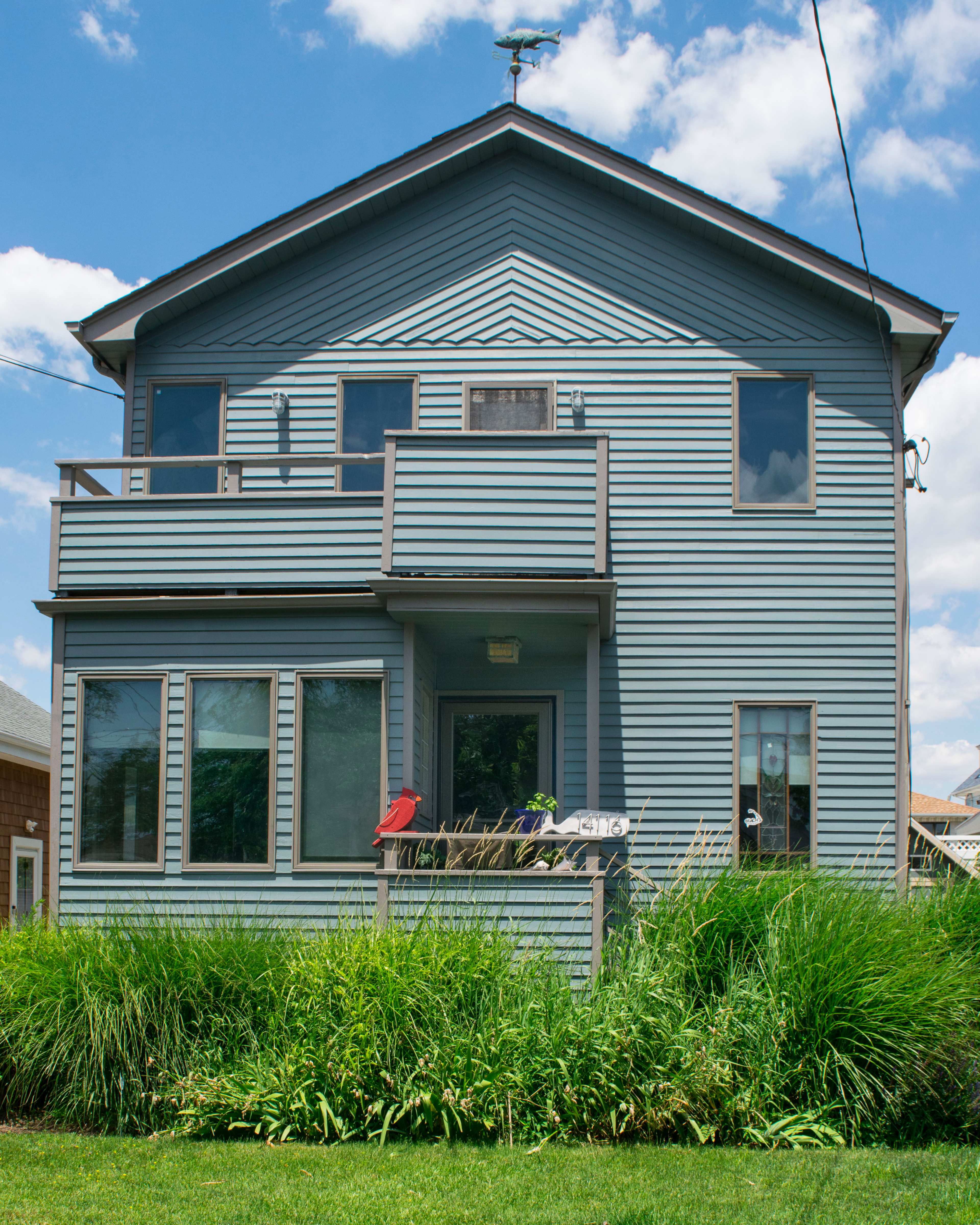 The image shows a two-story blue house with a balcony and several windows, surrounded by green grass and shrubs.
