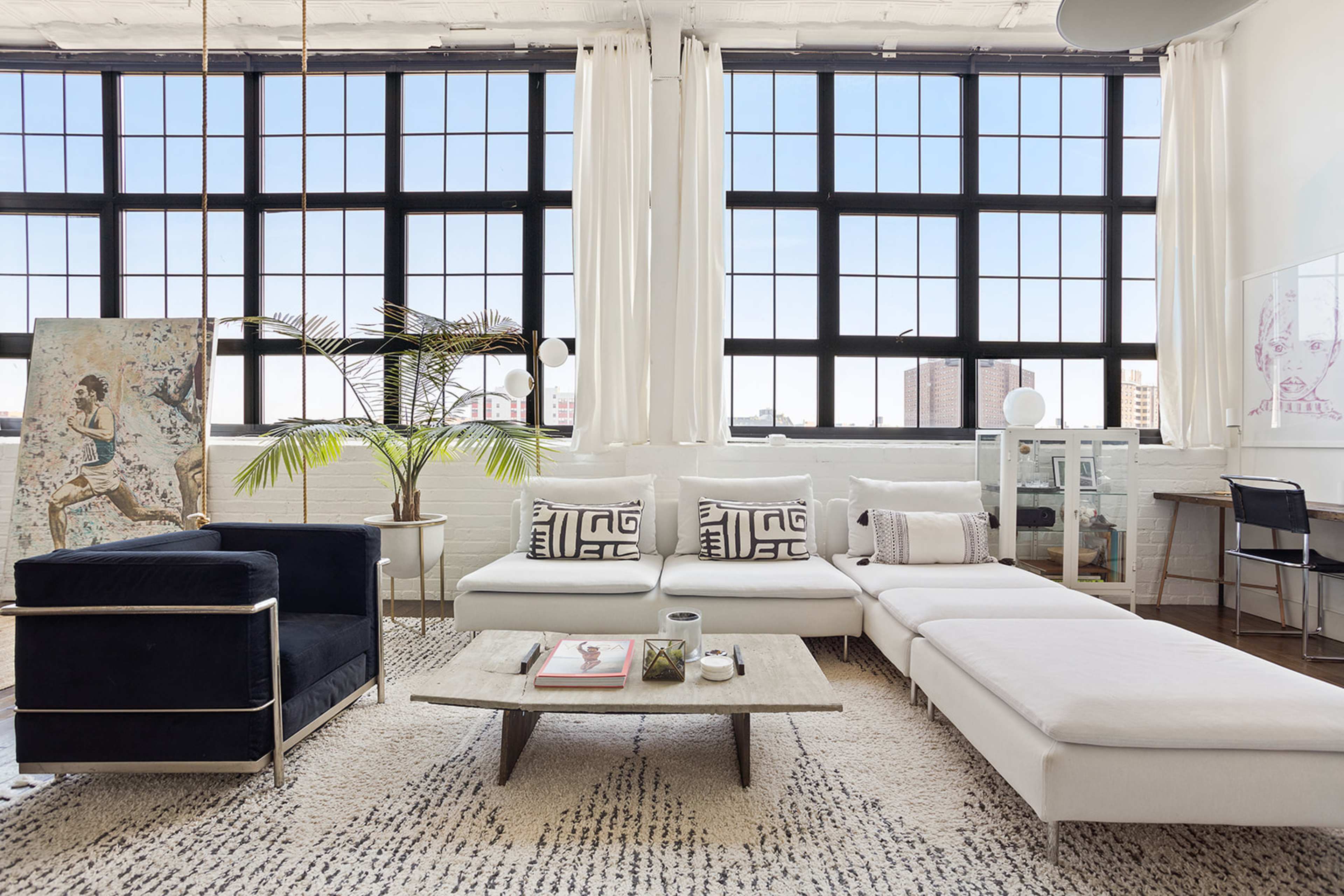 A modern living room with large windows, a white sectional sofa, a black armchair, a coffee table, and a desk beside a potted plant.