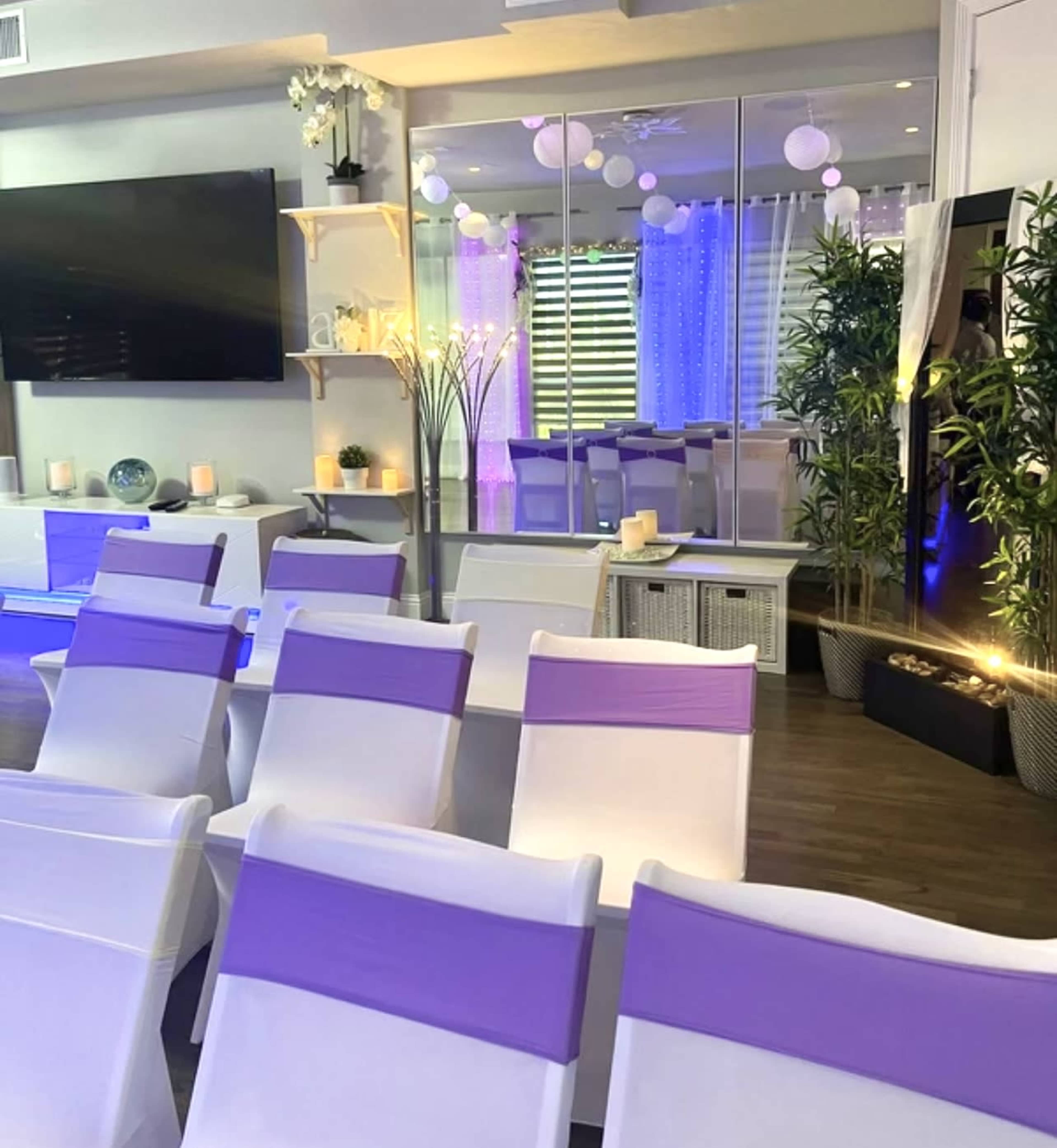 The image shows a modern event space featuring white chairs with purple sashes arranged in rows, a large TV screen, decorative plants, and ambient lighting.