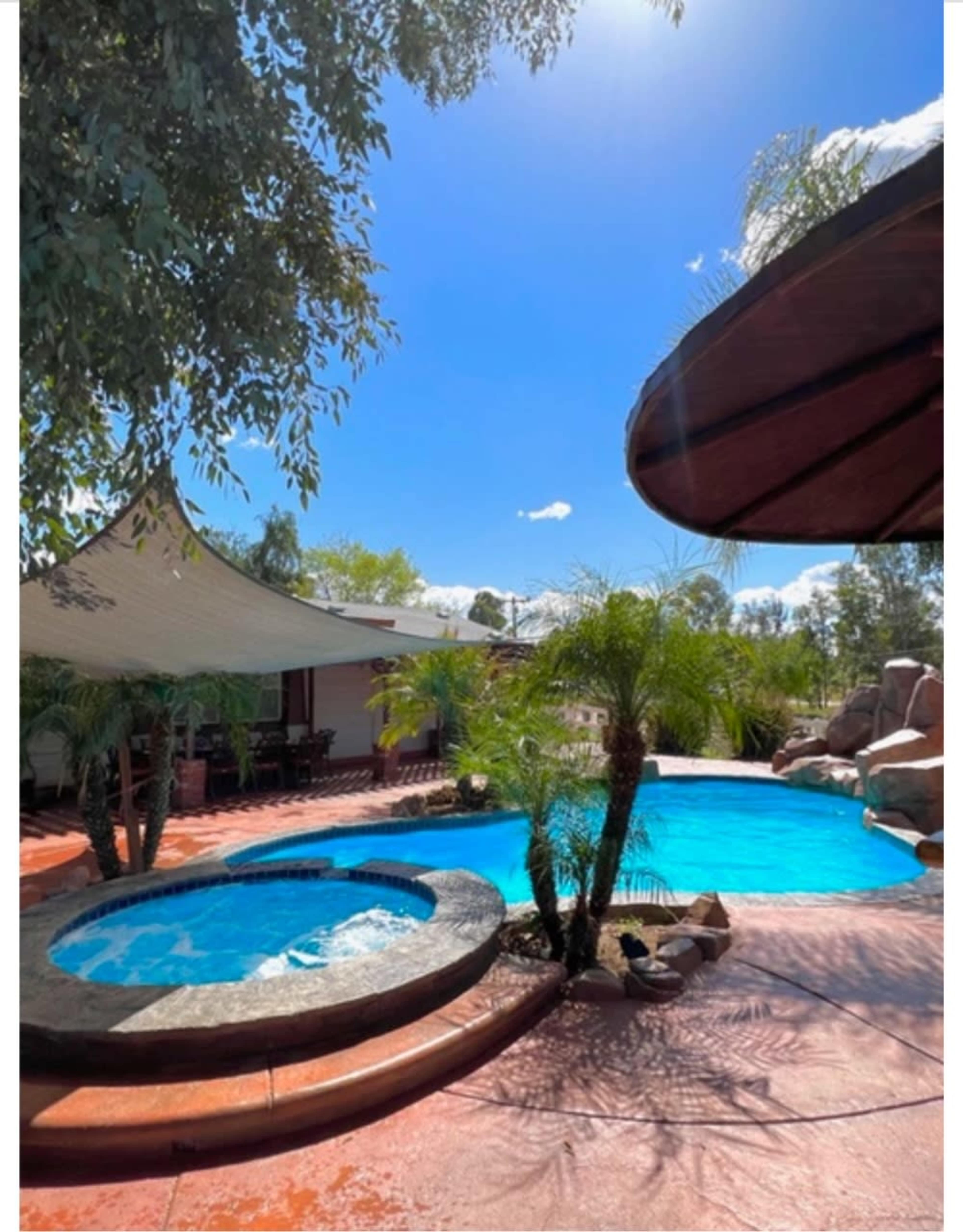 The image shows a landscaped backyard with a swimming pool, a hot tub, and tropical plants under a clear blue sky.