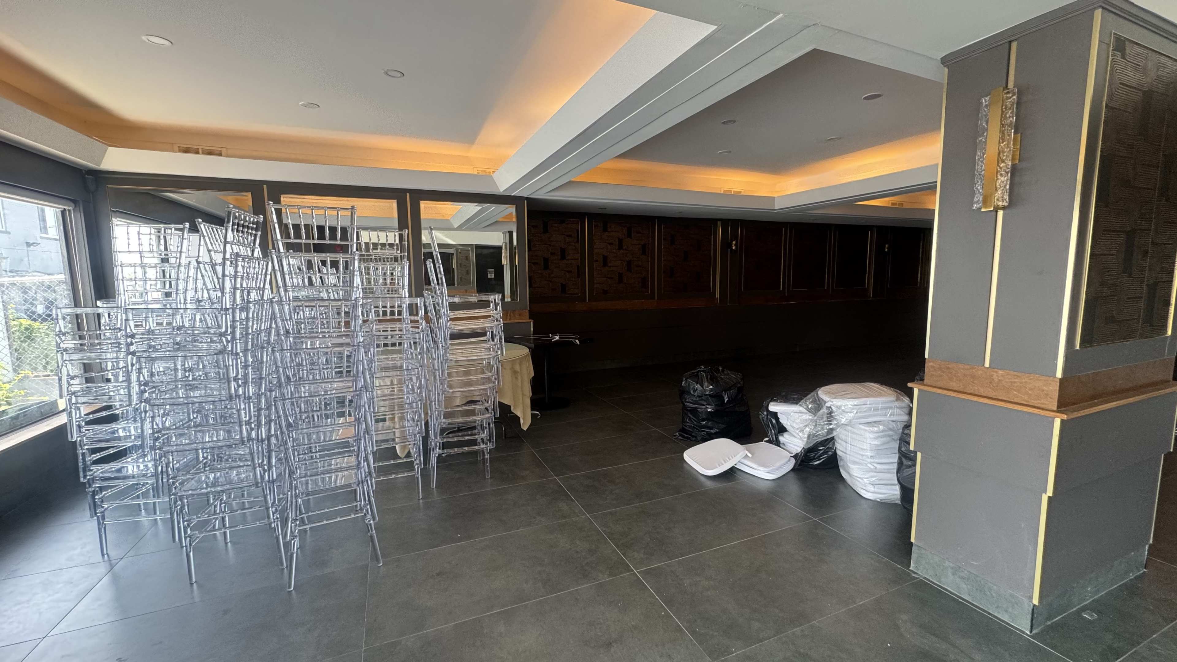 The image shows a partially empty indoor space with stacked transparent chairs on one side and piles of plastic bags and white chairs on the other.