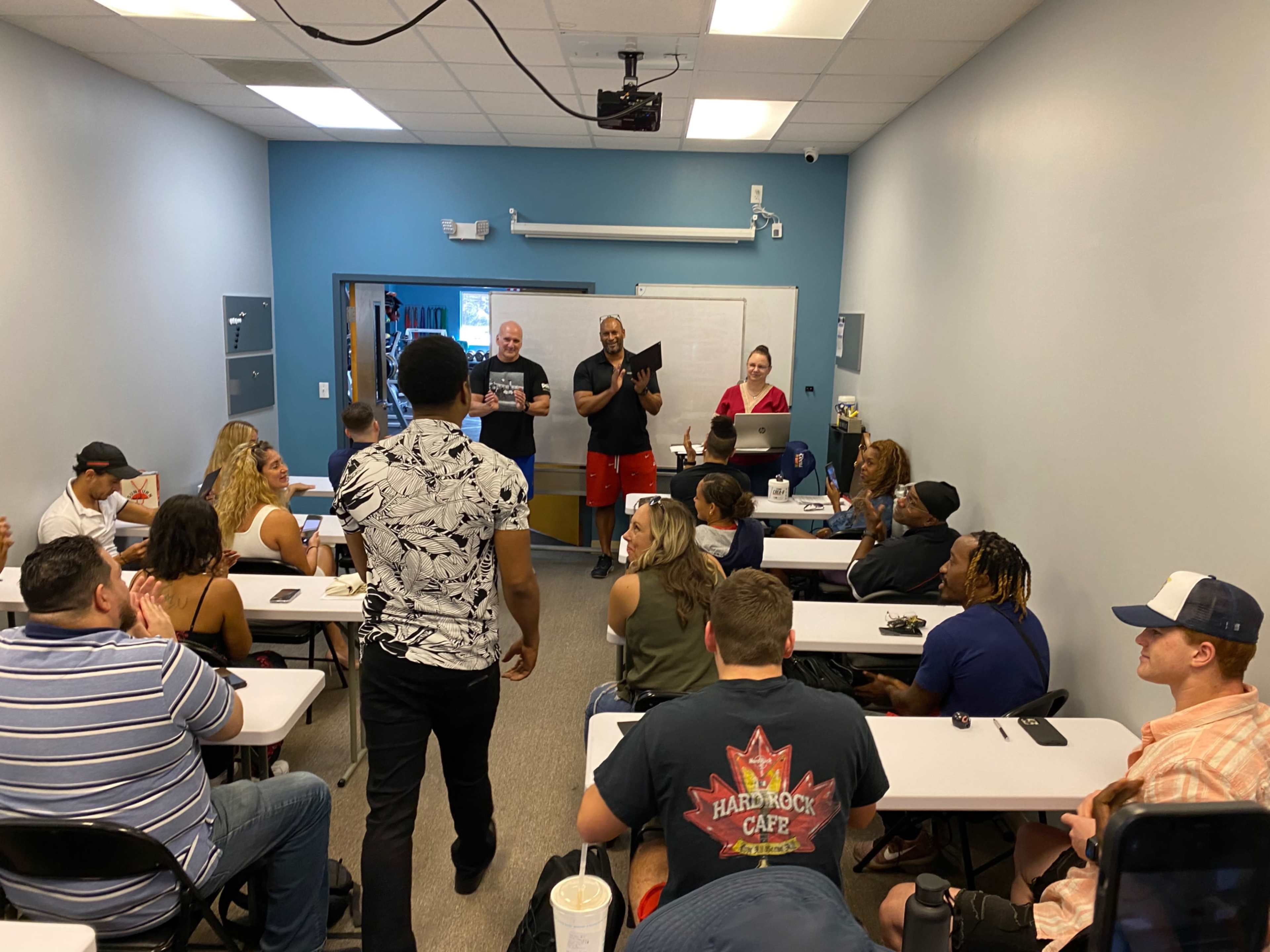 A group of people are seated at desks in a classroom, while three individuals stand at the front, engaging with the audience.
