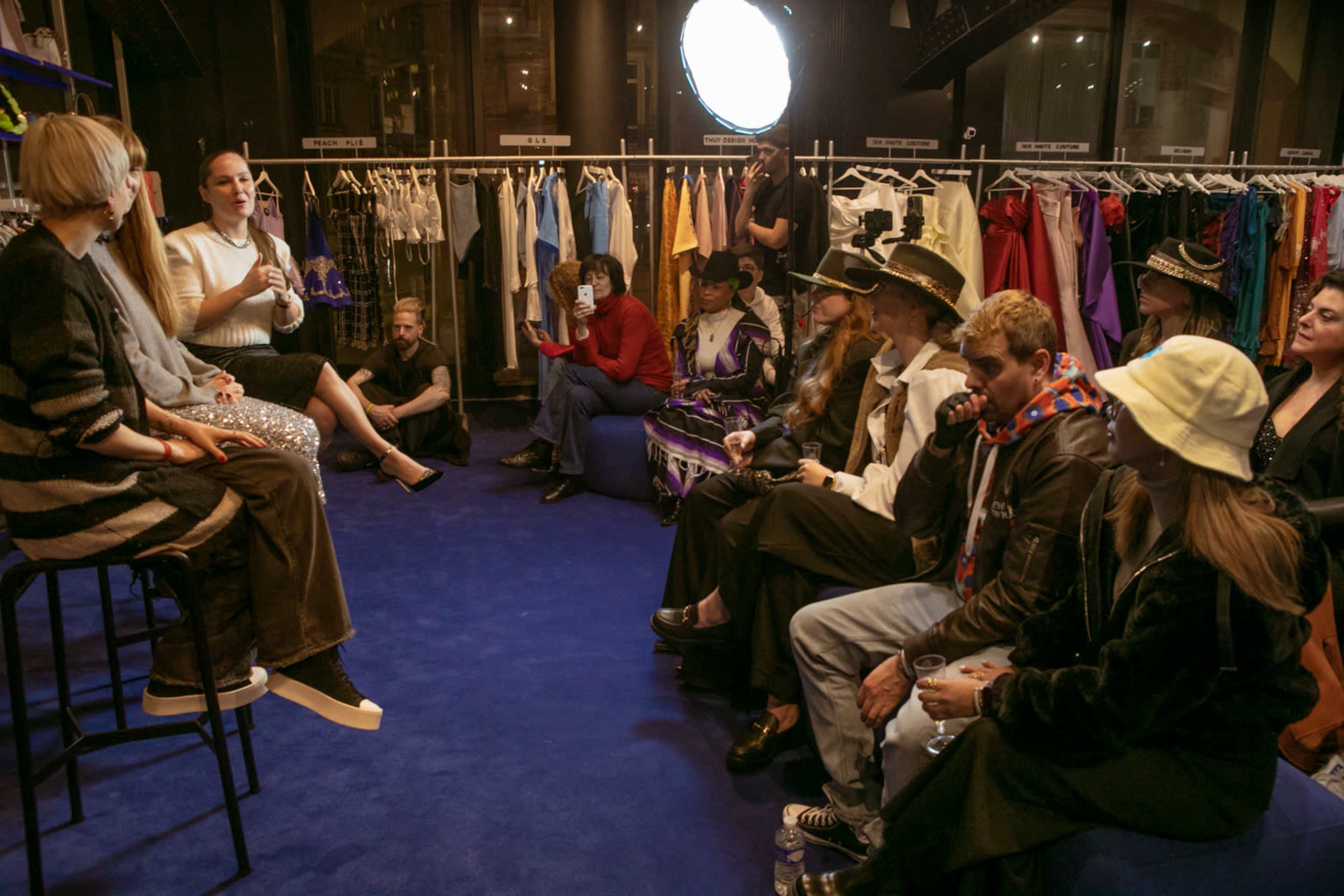 A group of people is seated on couches and stools in a clothing store, while a speaker discusses fashion-related topics amidst hanging garments.