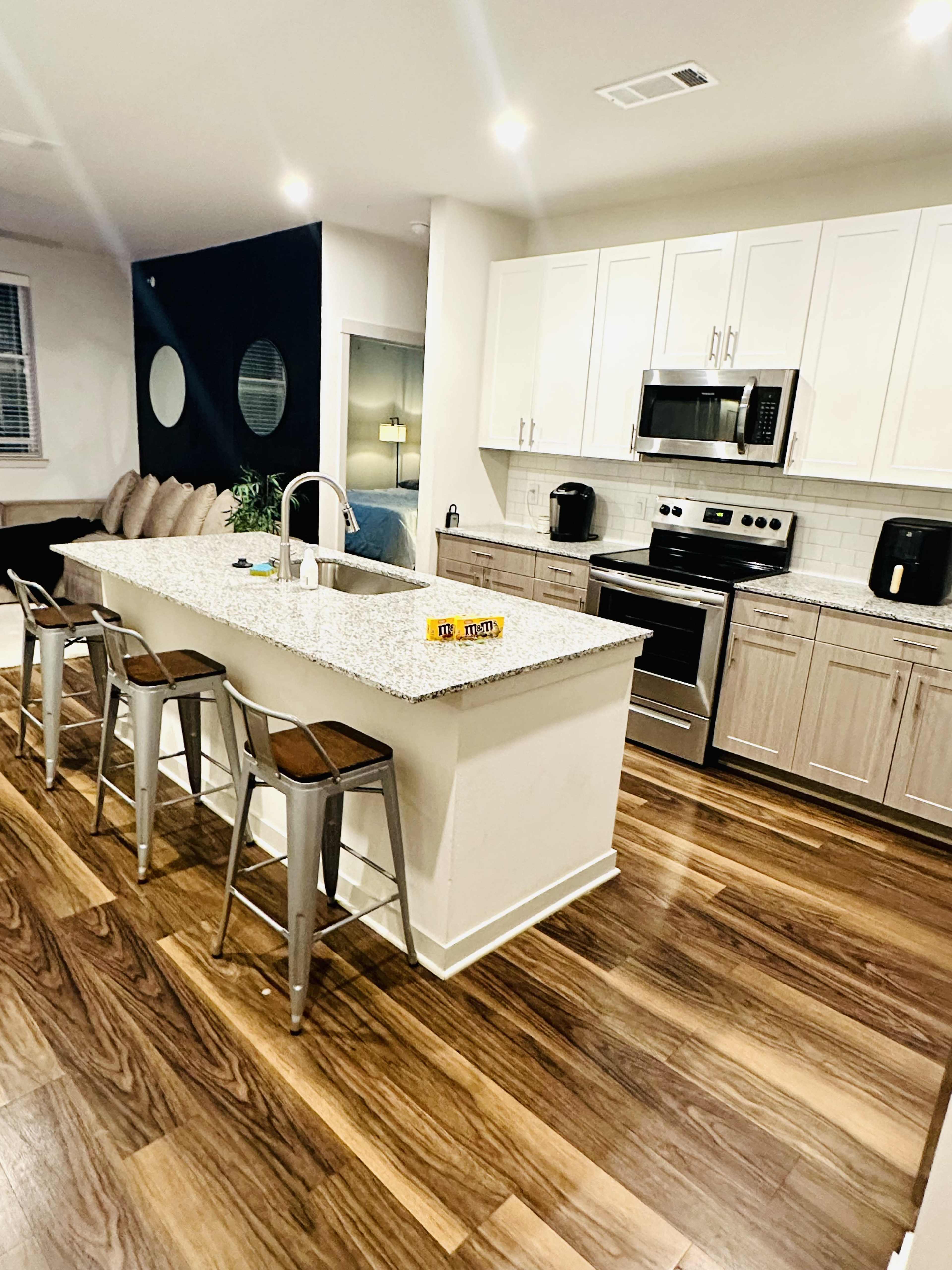 The image shows a modern kitchen with white cabinets, stainless steel appliances, a large island with four bar stools, and hardwood flooring.