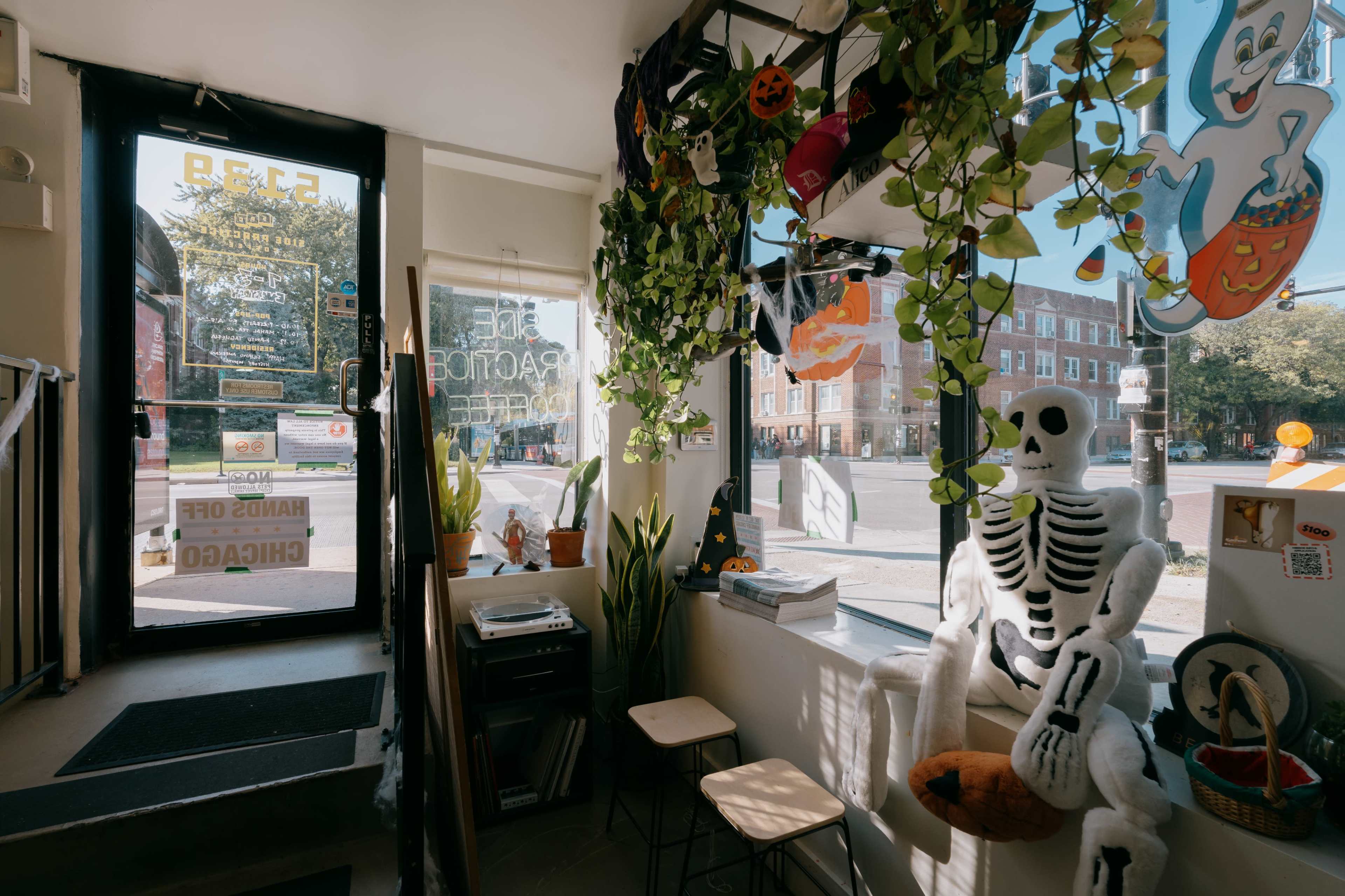 The image shows a cozy shop interior with Halloween decorations, including a skeleton and pumpkins, as well as large windows letting in natural light.