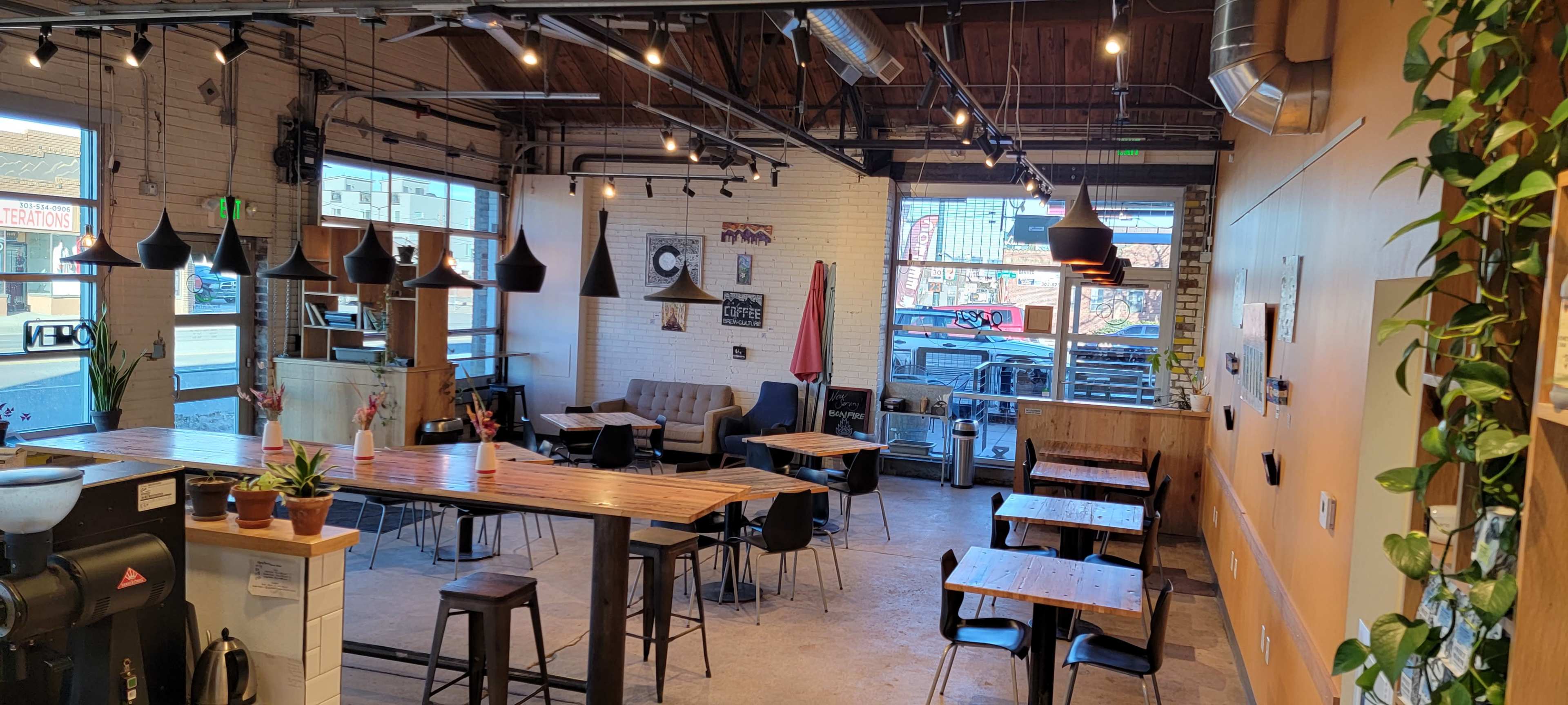 A spacious cafe interior with wooden tables, black chairs, and large windows allowing natural light to enter.