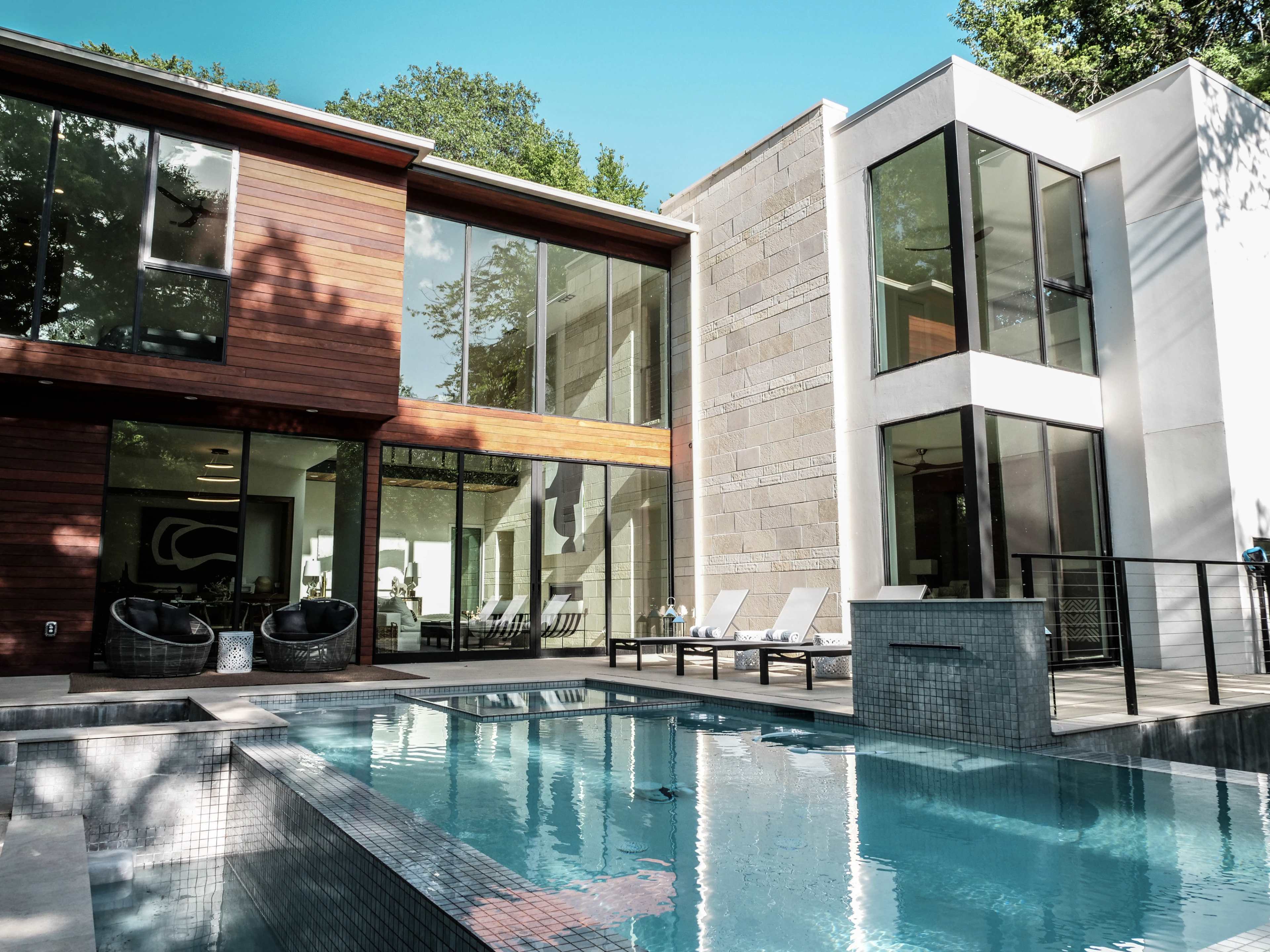 A modern house with large windows overlooks a rectangular swimming pool surrounded by lounge chairs.