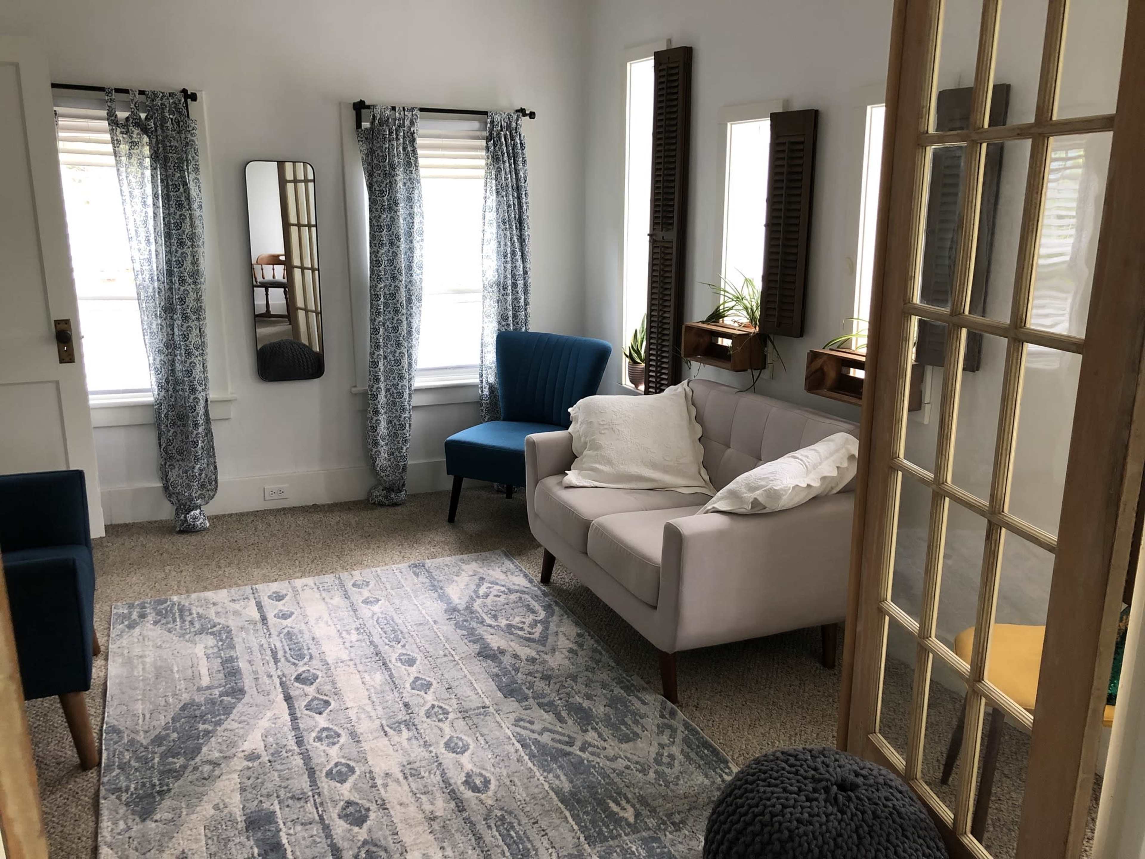 The room features a light-colored sofa, two blue chairs, a patterned rug, and large windows with curtains, all arranged around a set of plants and mirrors.
