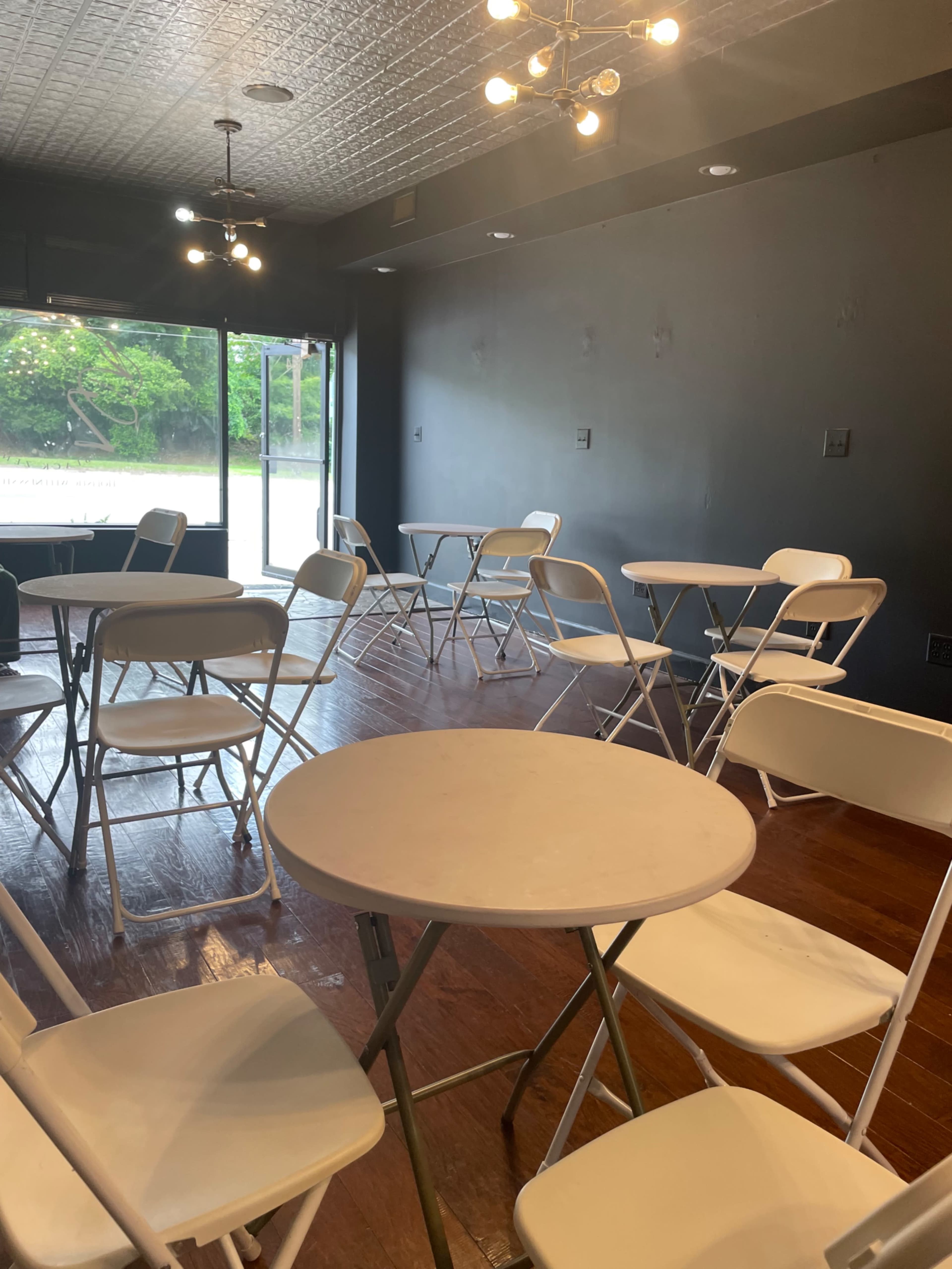 A room with several white folding tables and chairs arranged on a hardwood floor, featuring a large window overlooking an outdoor area.