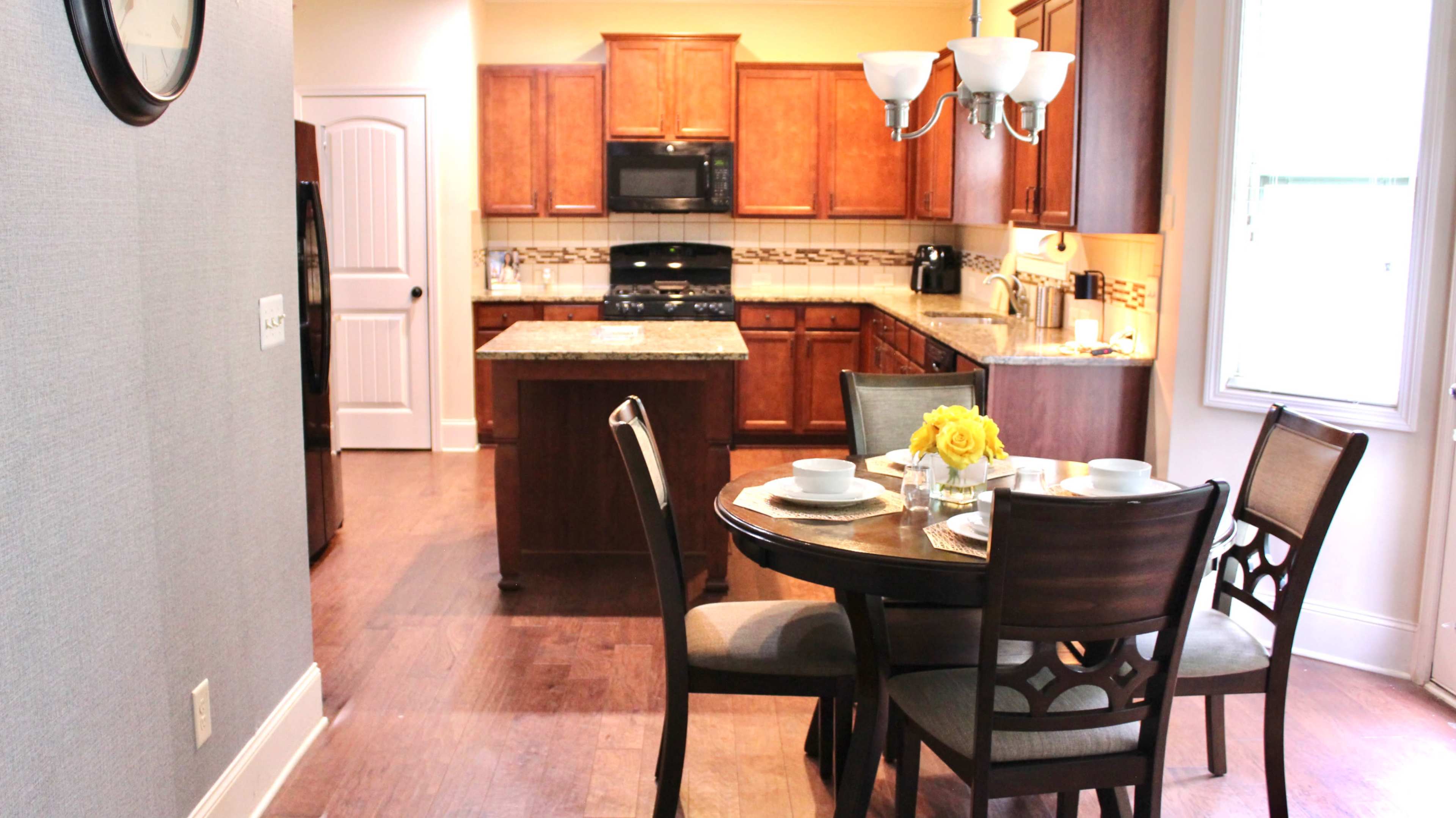 The image shows a modern kitchen and dining area, featuring wooden cabinets, a granite countertop, and a round dining table set for four with a vase of yellow flowers.