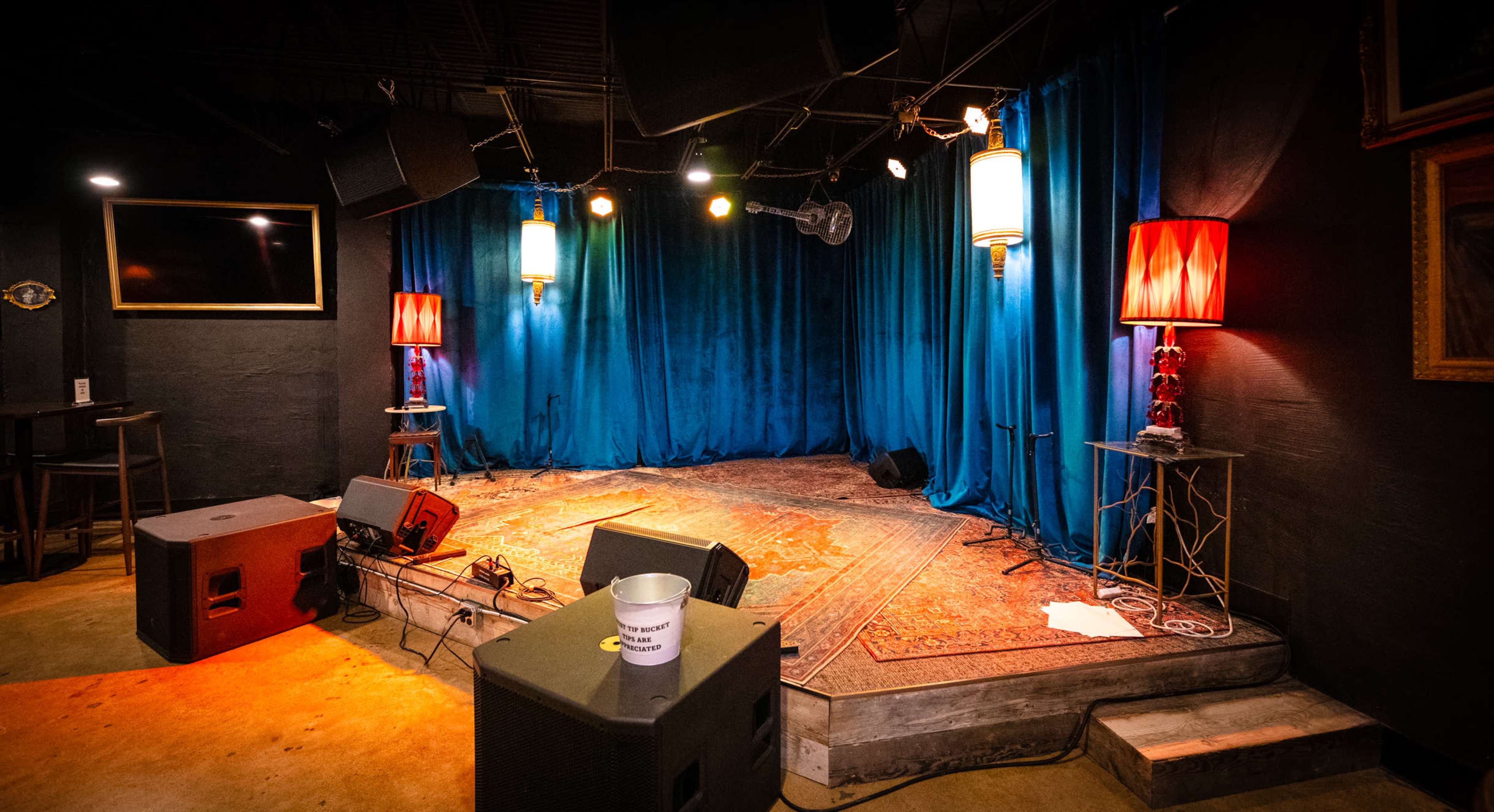 The image shows a small stage area with blue curtains, two red lamps, and sound equipment, set up for performances in a low-lit venue.