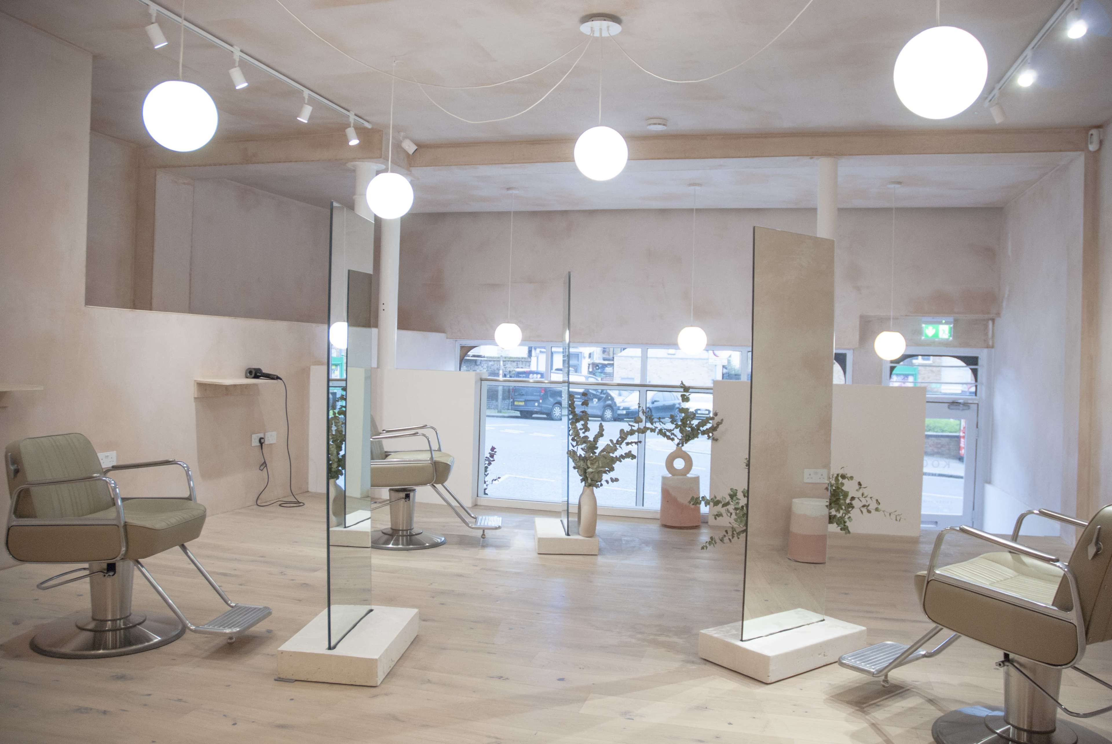 The image shows a modern hair salon featuring two styling chairs, tall mirrors, and minimalist decor with soft lighting.