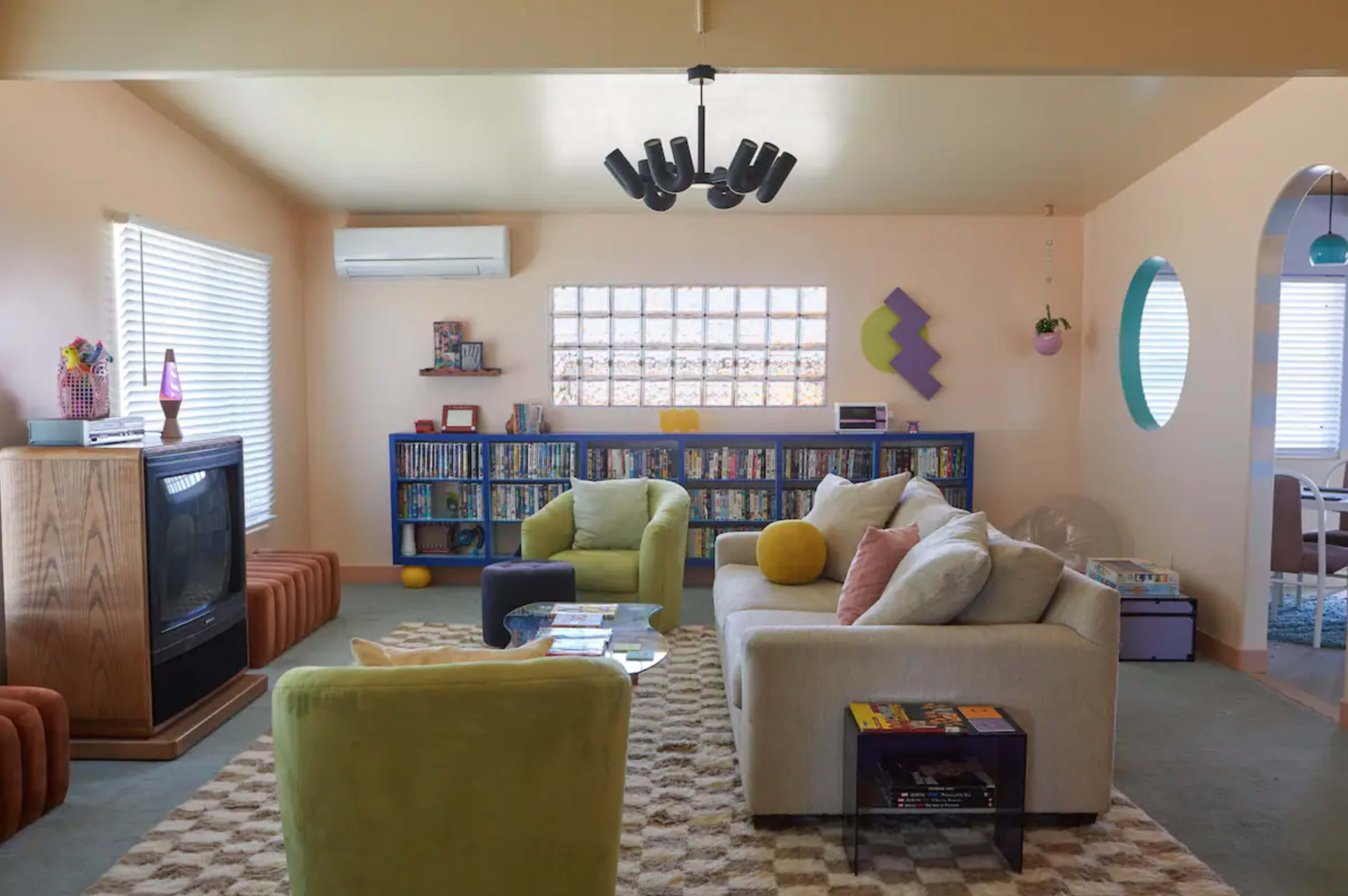 The living room features a light-colored sofa and chairs, a bookshelf filled with DVDs, and large windows allowing natural light to enter.