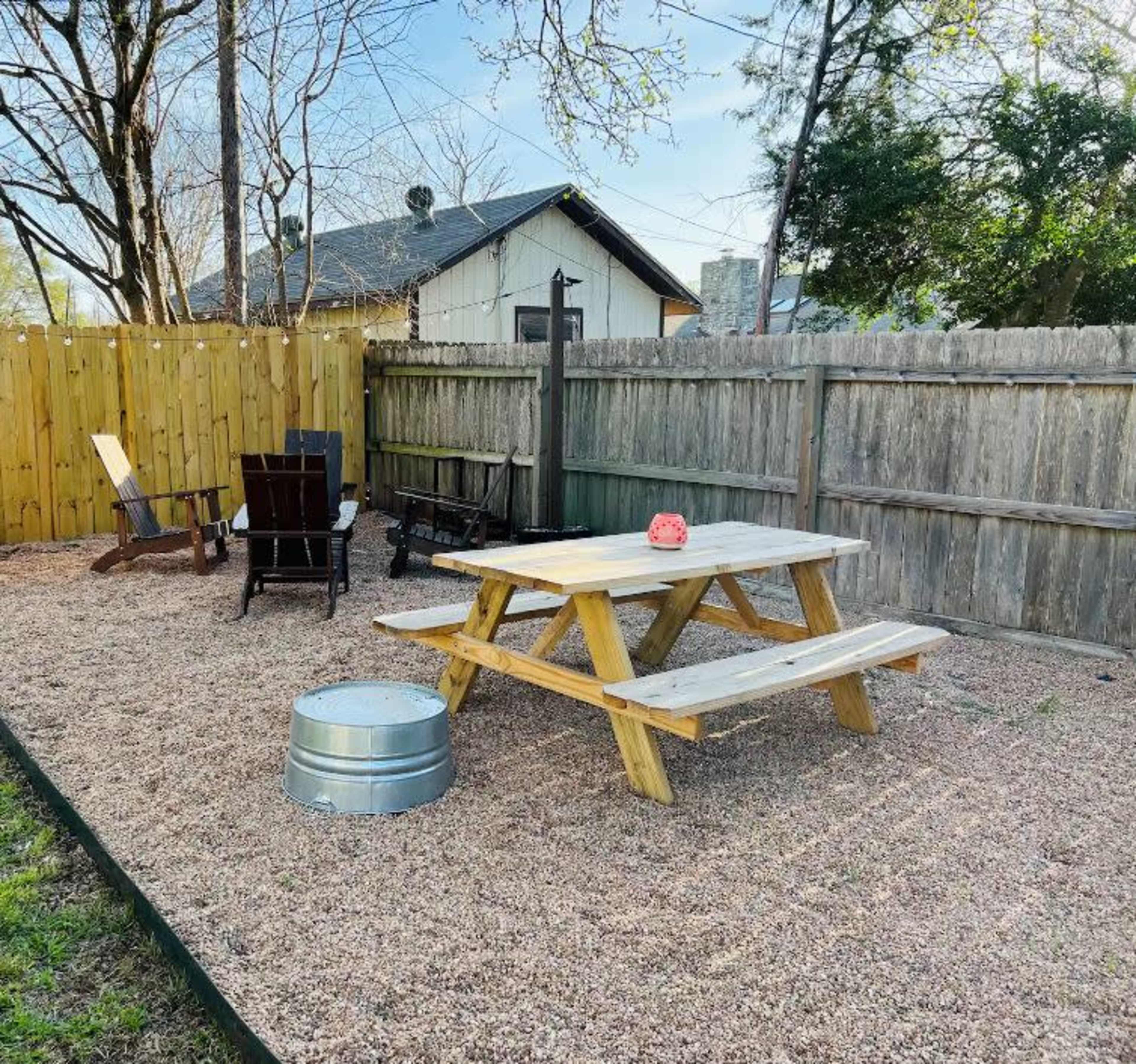The image shows a gravel-covered outdoor space with a wooden picnic table, two black chairs, and a metal fire pit surrounded by a wooden fence and trees.