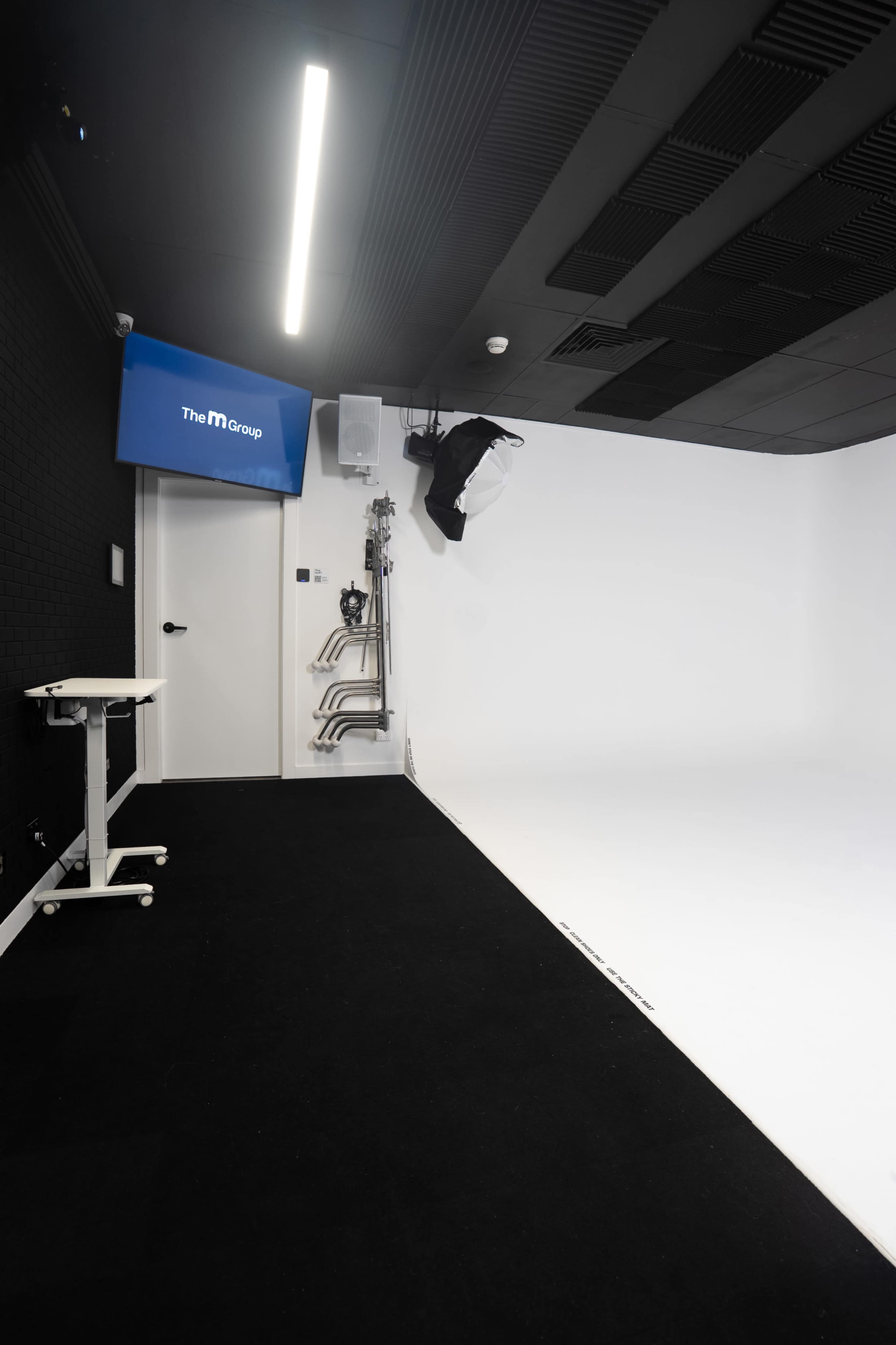 The image shows a minimalistic studio space with a white backdrop, black flooring, and a monitor displaying "The M Group" mounted on the wall.