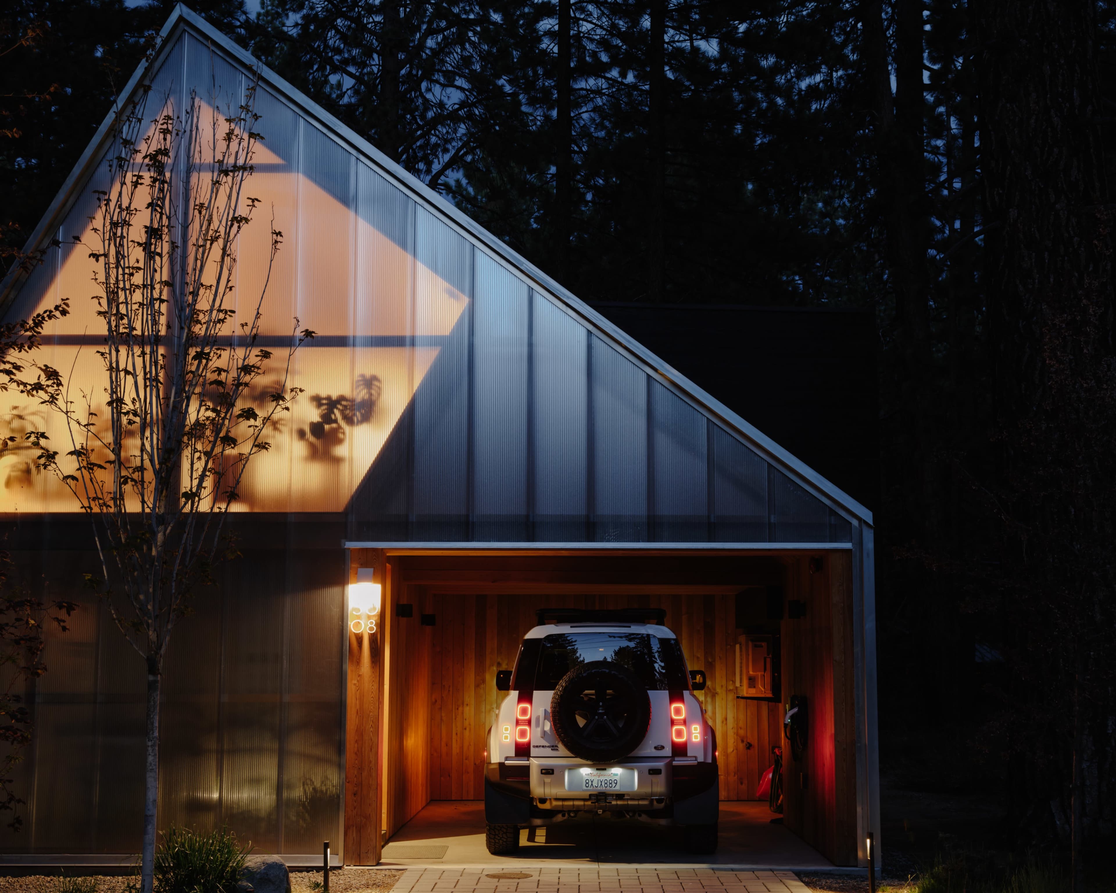 A vehicle is parked inside a carport of a modern house illuminated by warm lights at dusk.