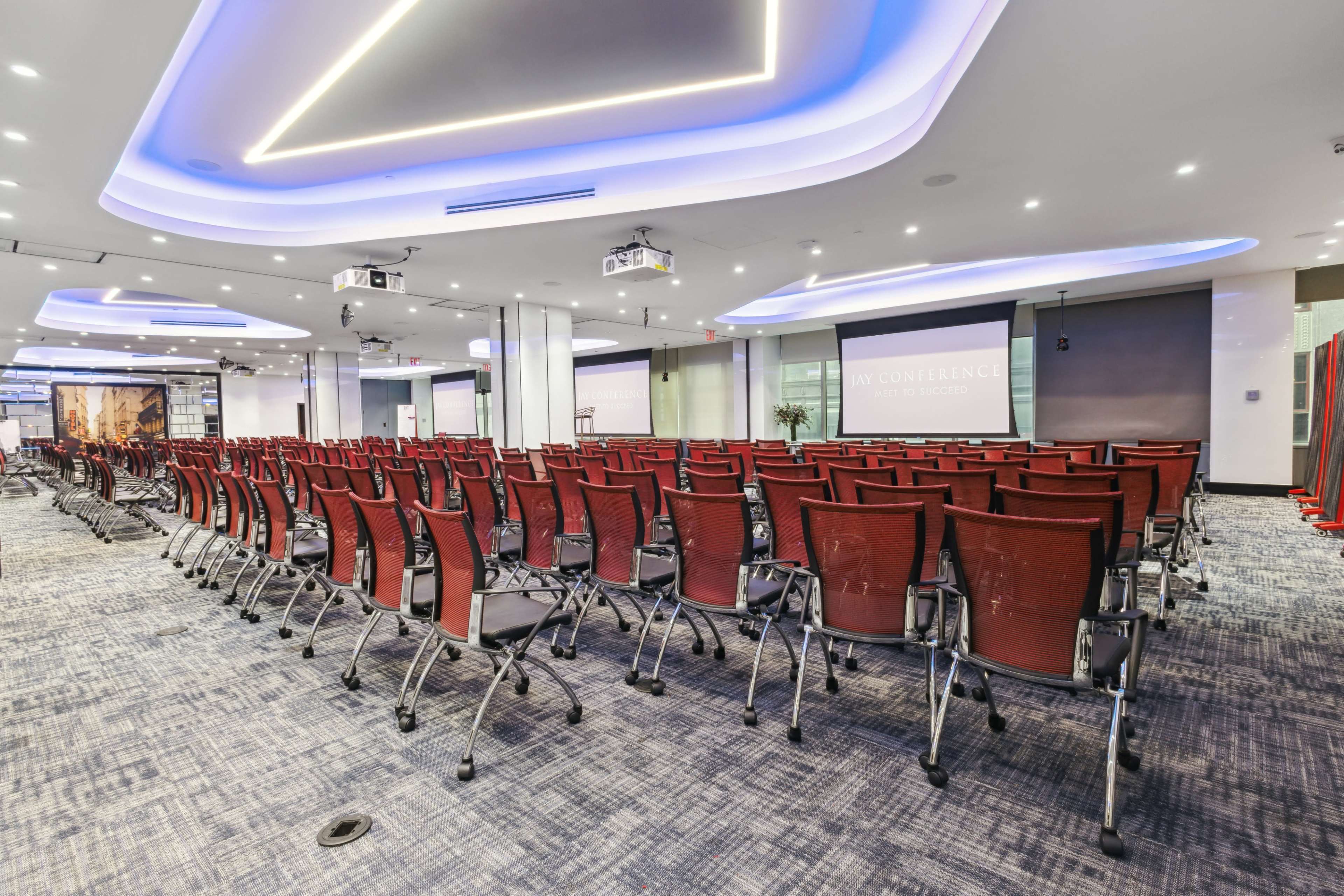 A spacious conference room is set up with rows of red chairs facing a large screen and projector, surrounded by modern lighting and decor.