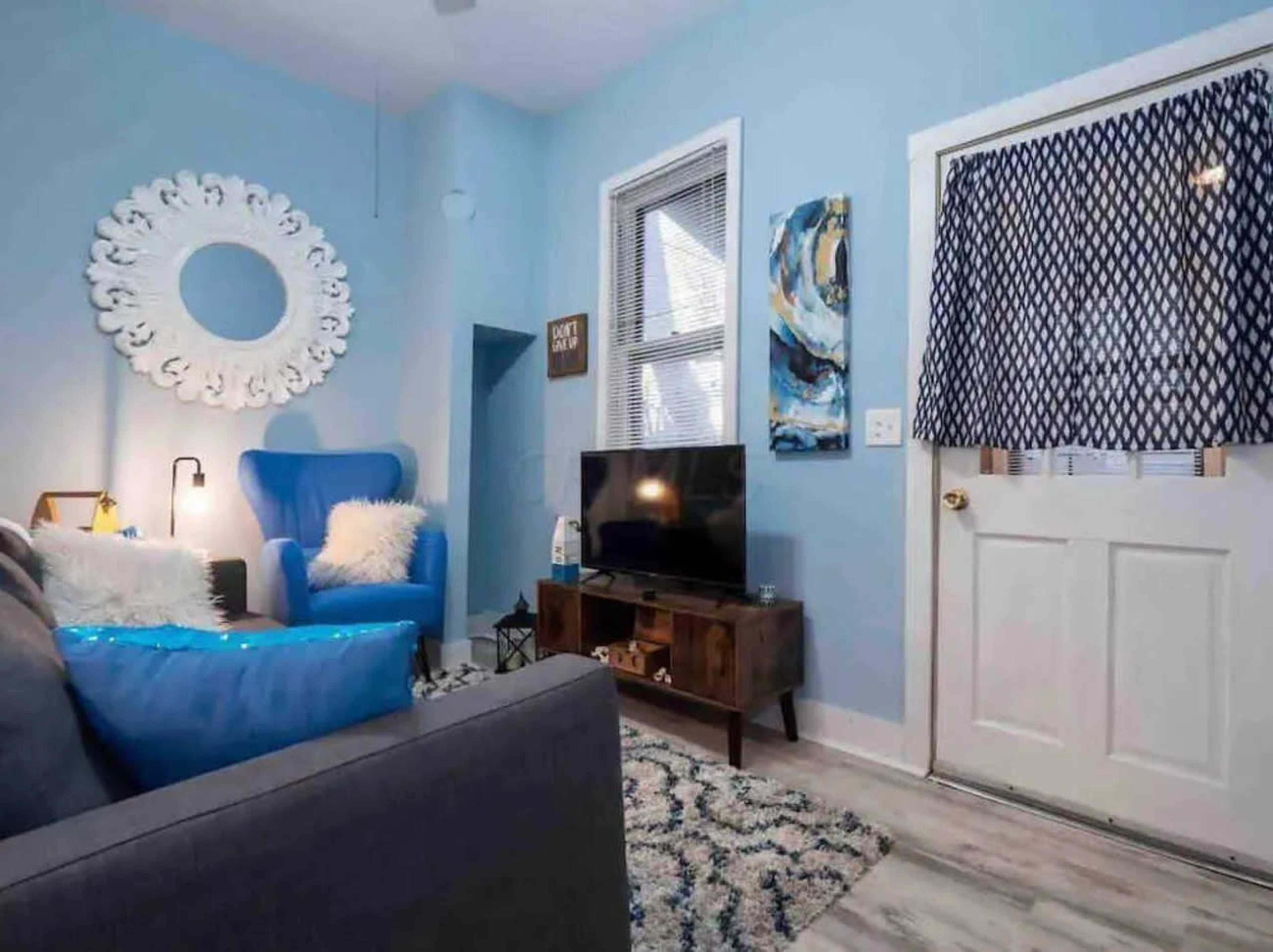 The image shows a small living room with a blue accent wall, a couch, a blue chair, a TV on a wooden stand, and a door with a patterned curtain.
