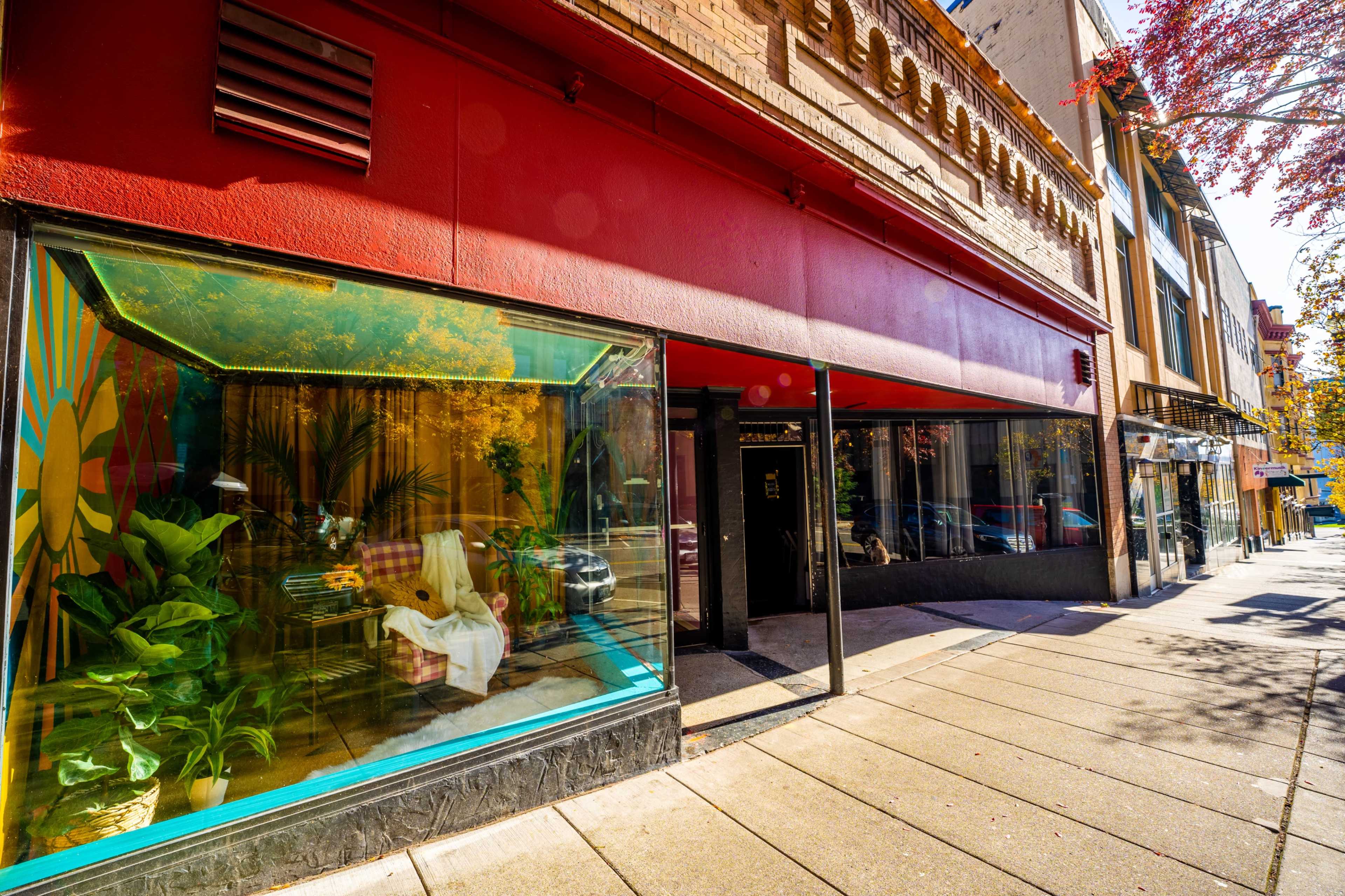 The image shows a vibrant storefront with large glass windows displaying plants and seating, set against a red exterior wall.