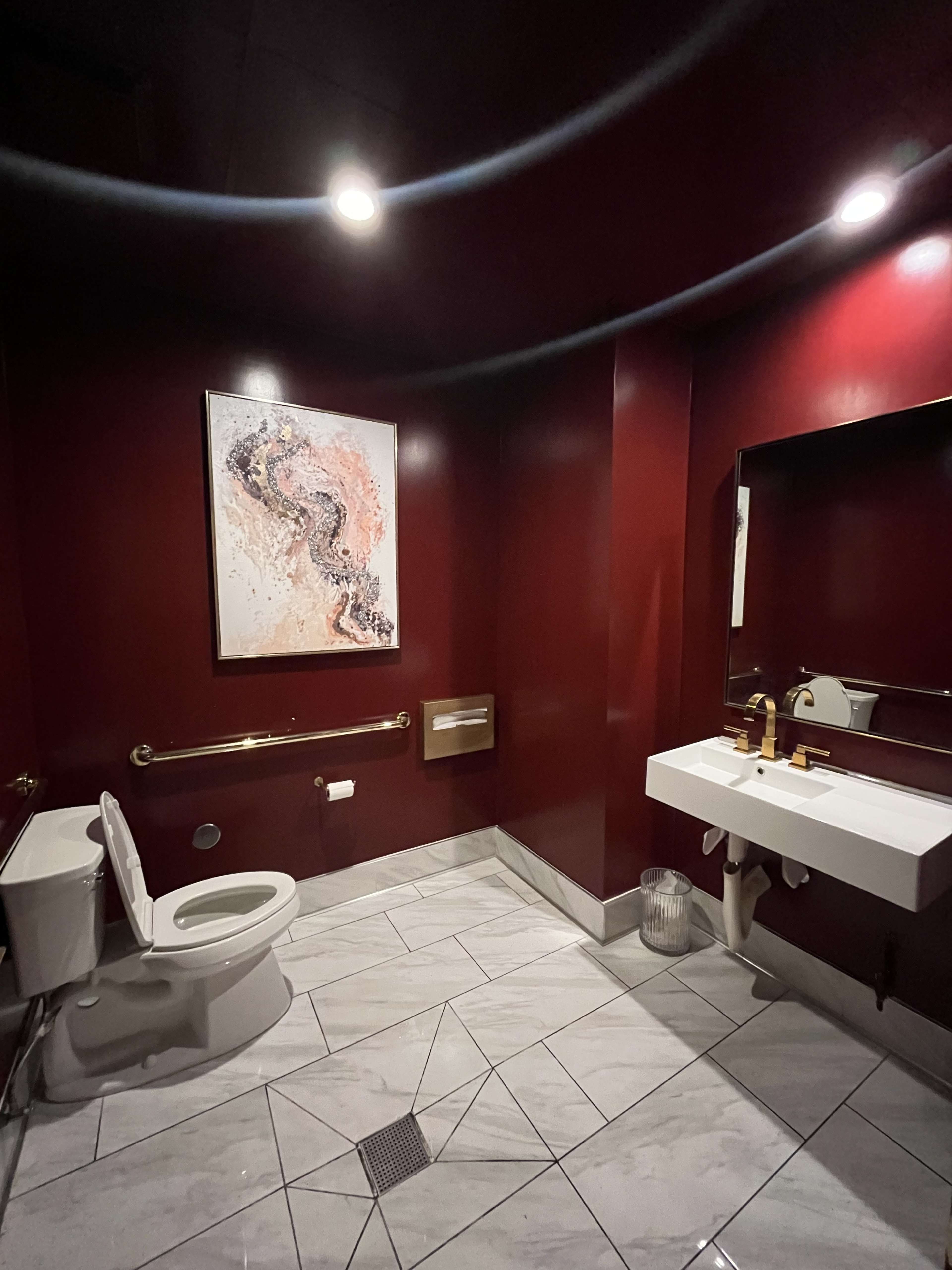 A bathroom with a toilet, a modern sink, a large mirror, and art on the wall, all set against burgundy walls and tiled flooring.